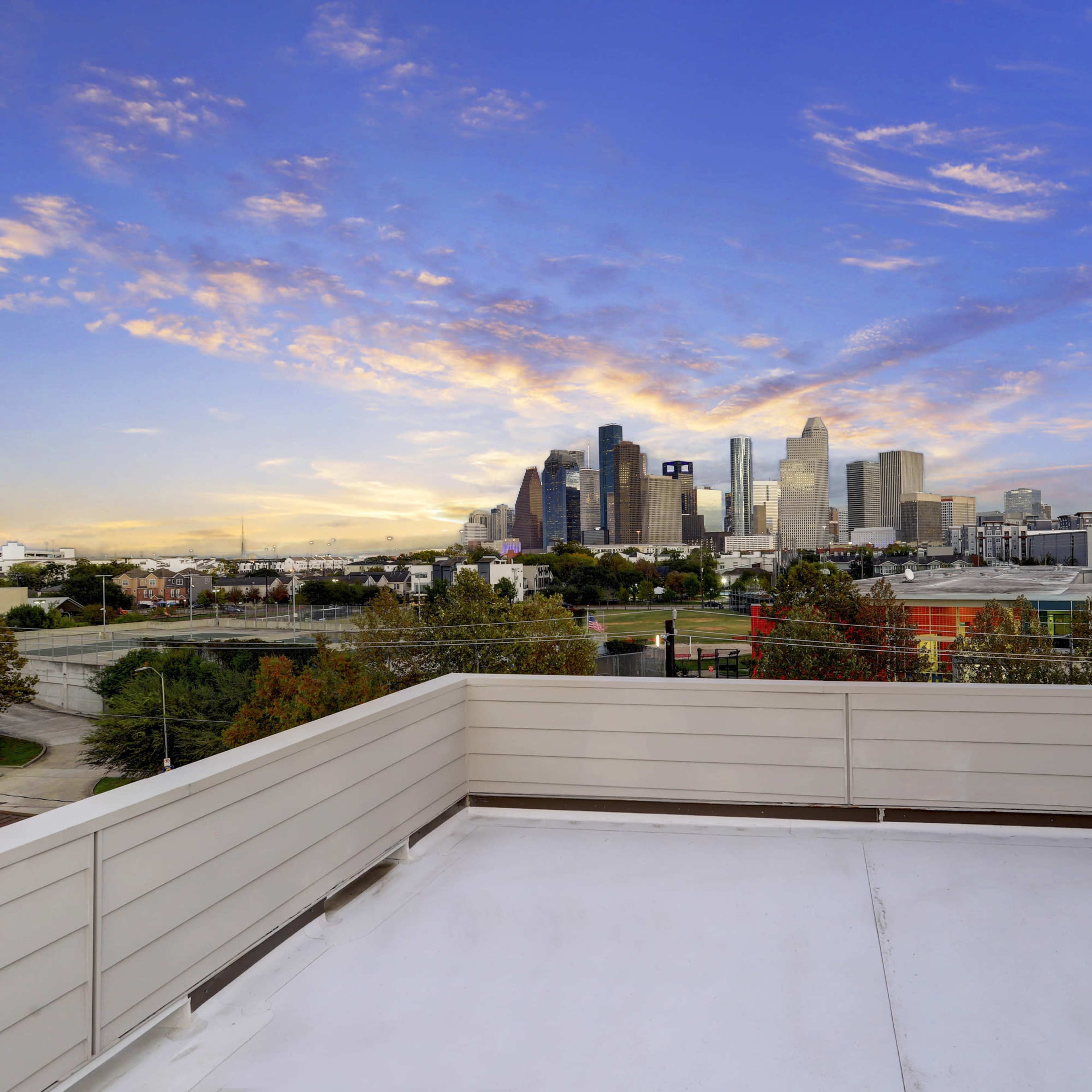 A rooftop terrace with a view of a city skyline during sunset, featuring various buildings and a colorful sky.
