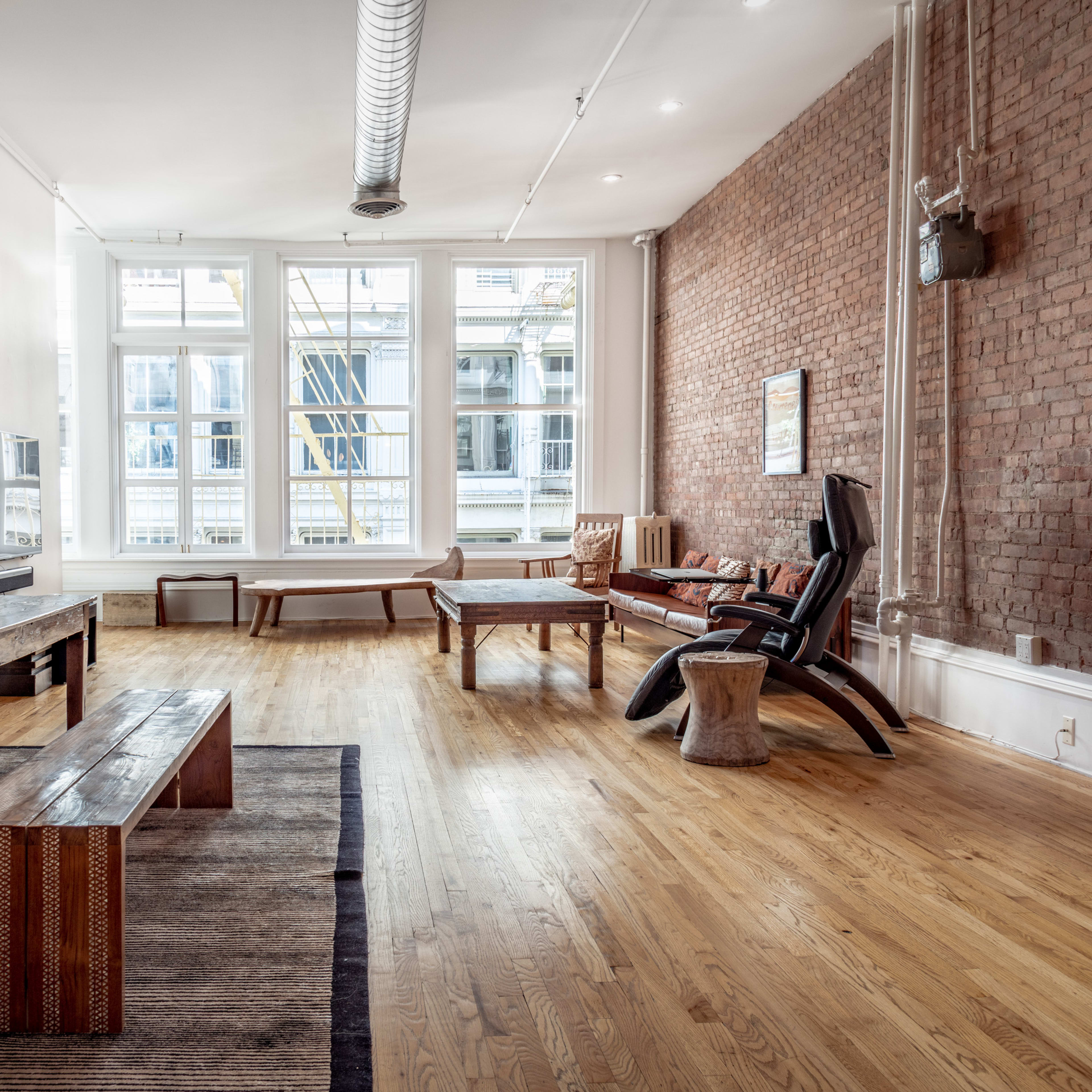A spacious, industrial-style living room with hardwood floors, exposed brick walls, large windows, and various wooden furniture pieces.