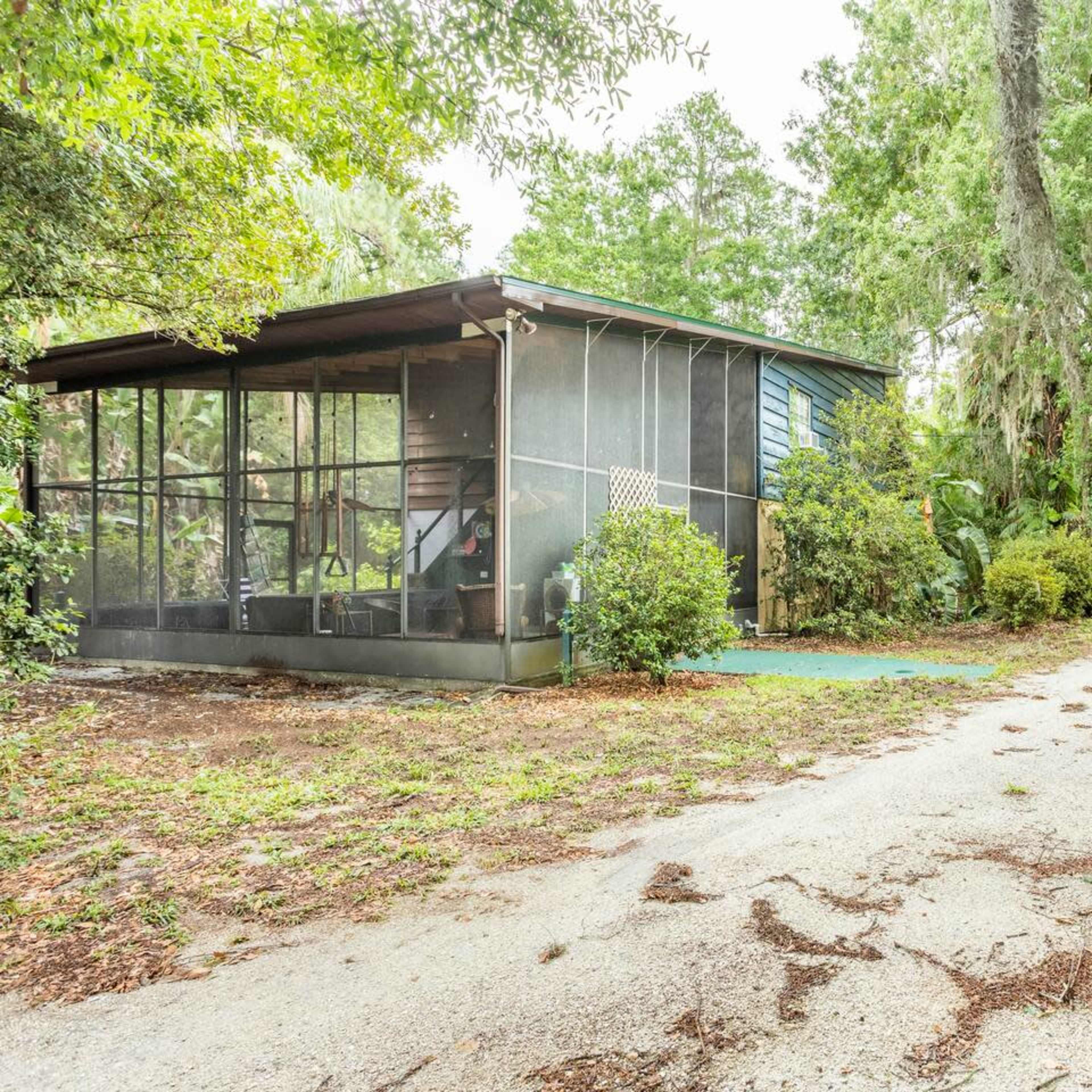 A gravel driveway leads to a small house surrounded by greenery, with a blue shed and recycling bin nearby.