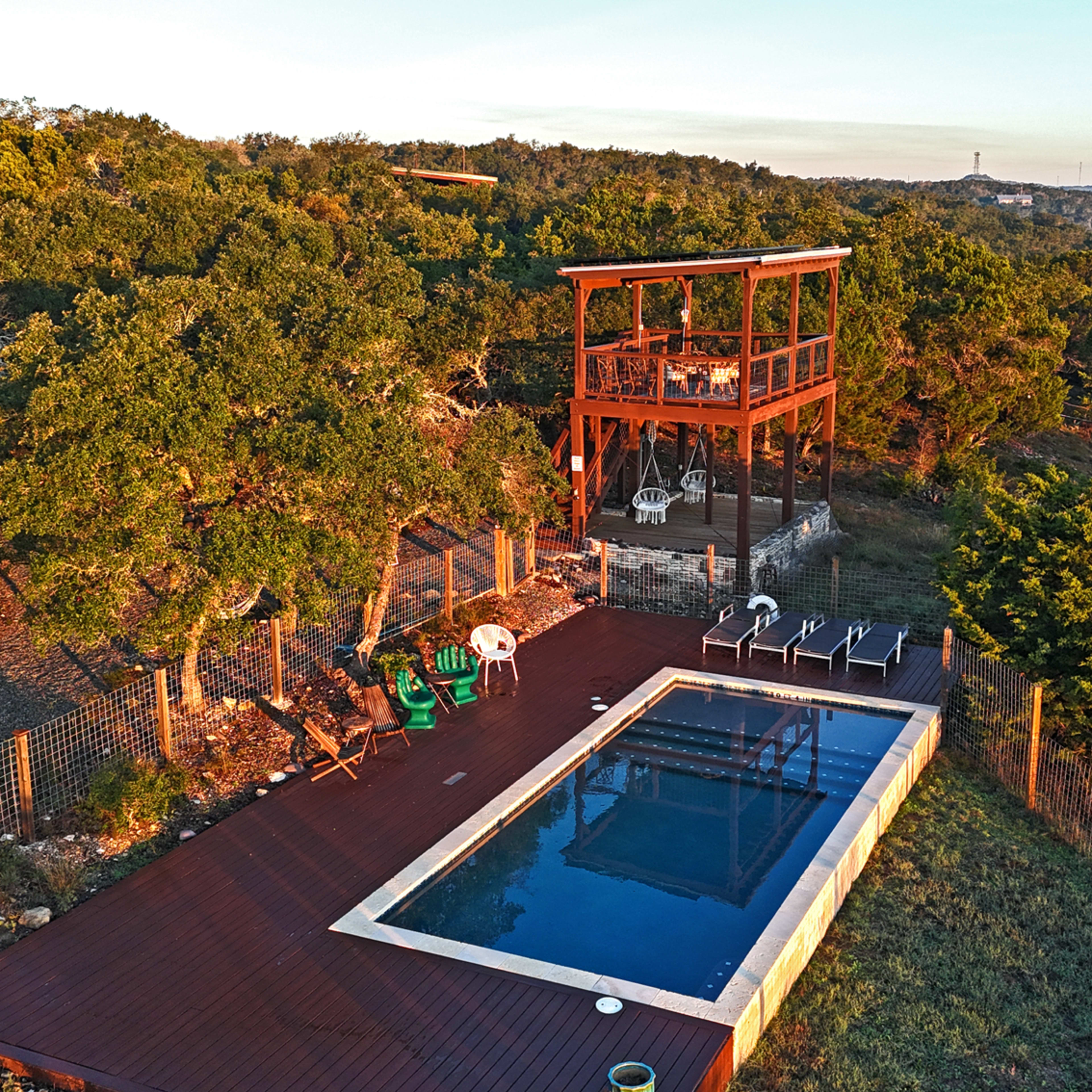 A wooden deck surrounding a rectangular swimming pool, located on a hillside amid trees, with a multi-level observation tower overlooking the area.