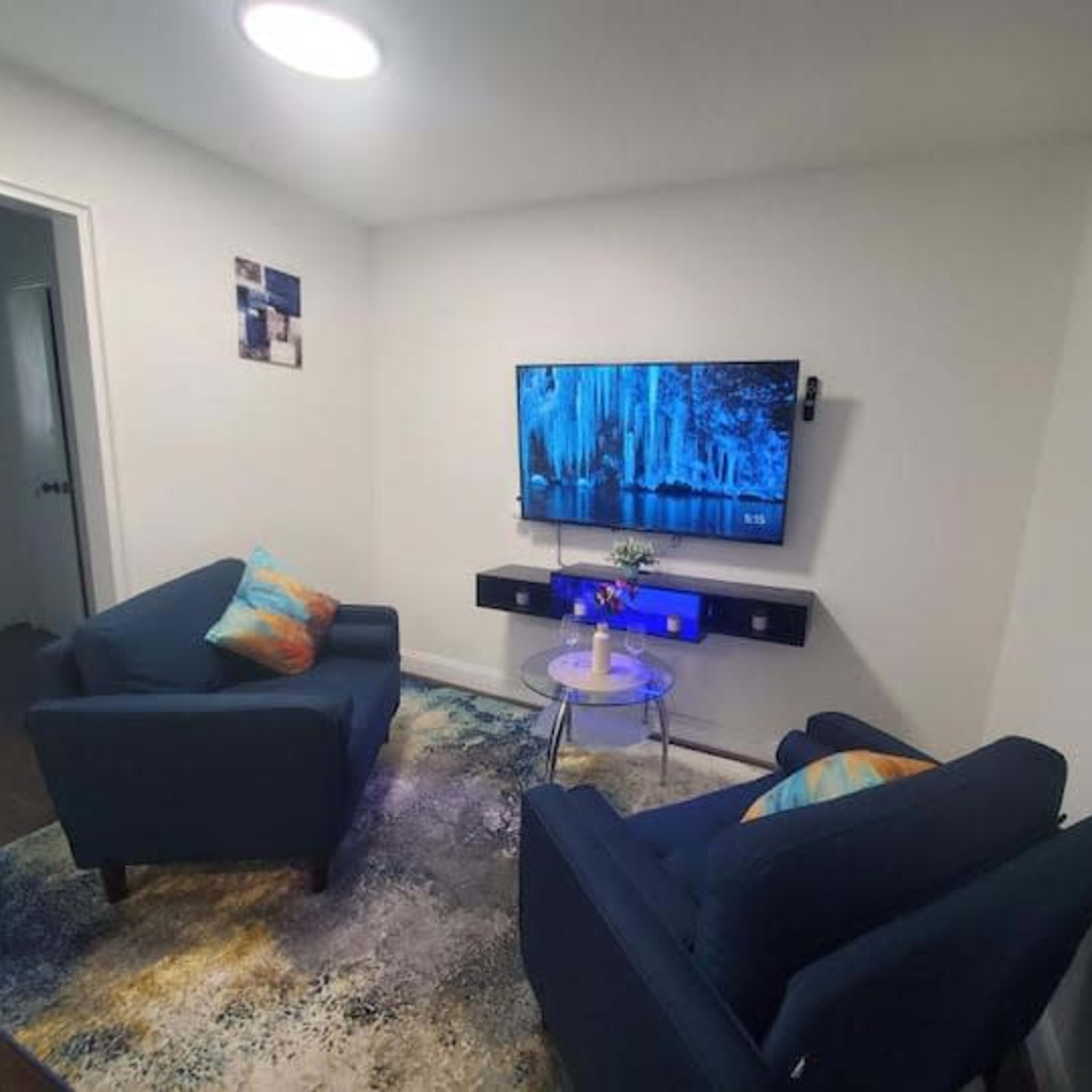 The room features two blue chairs facing a wall-mounted television, with a small table and a decorative rug on the floor.