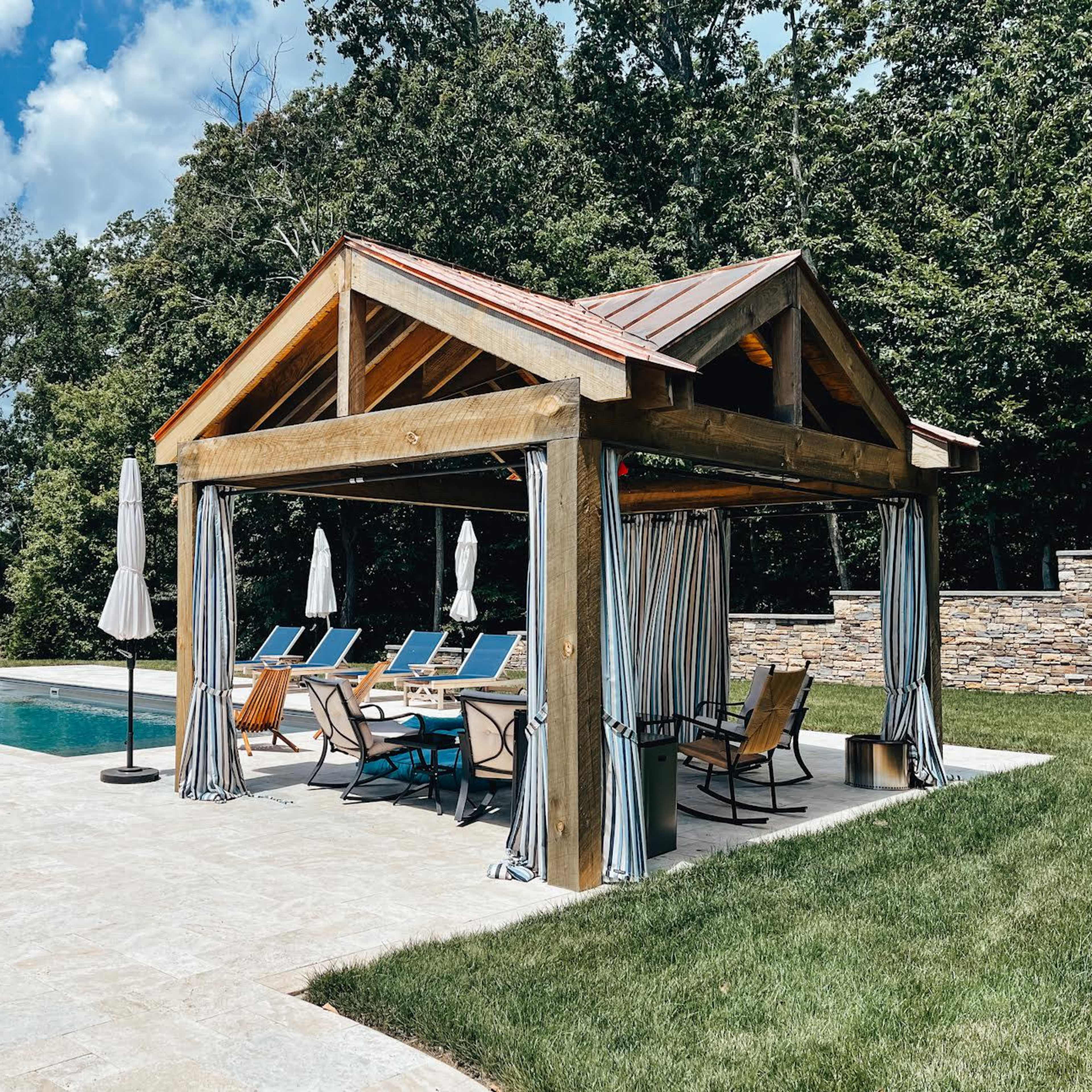 A wooden pavilion with a sloped roof and striped curtains shelters lounge chairs by a swimming pool surrounded by trees.