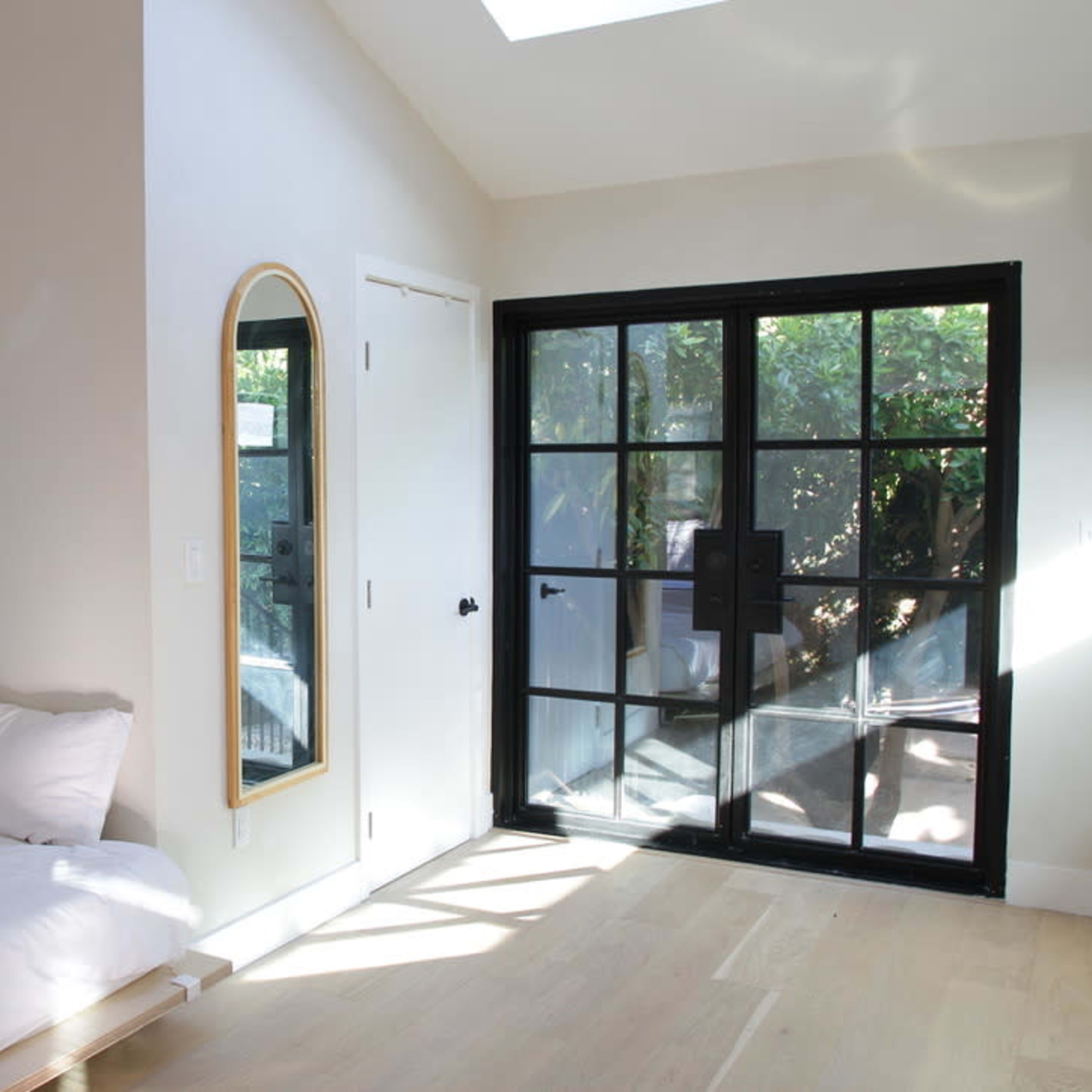 A small bedroom features a bed against one wall, a mirror, and a glass door leading to an outdoor area.