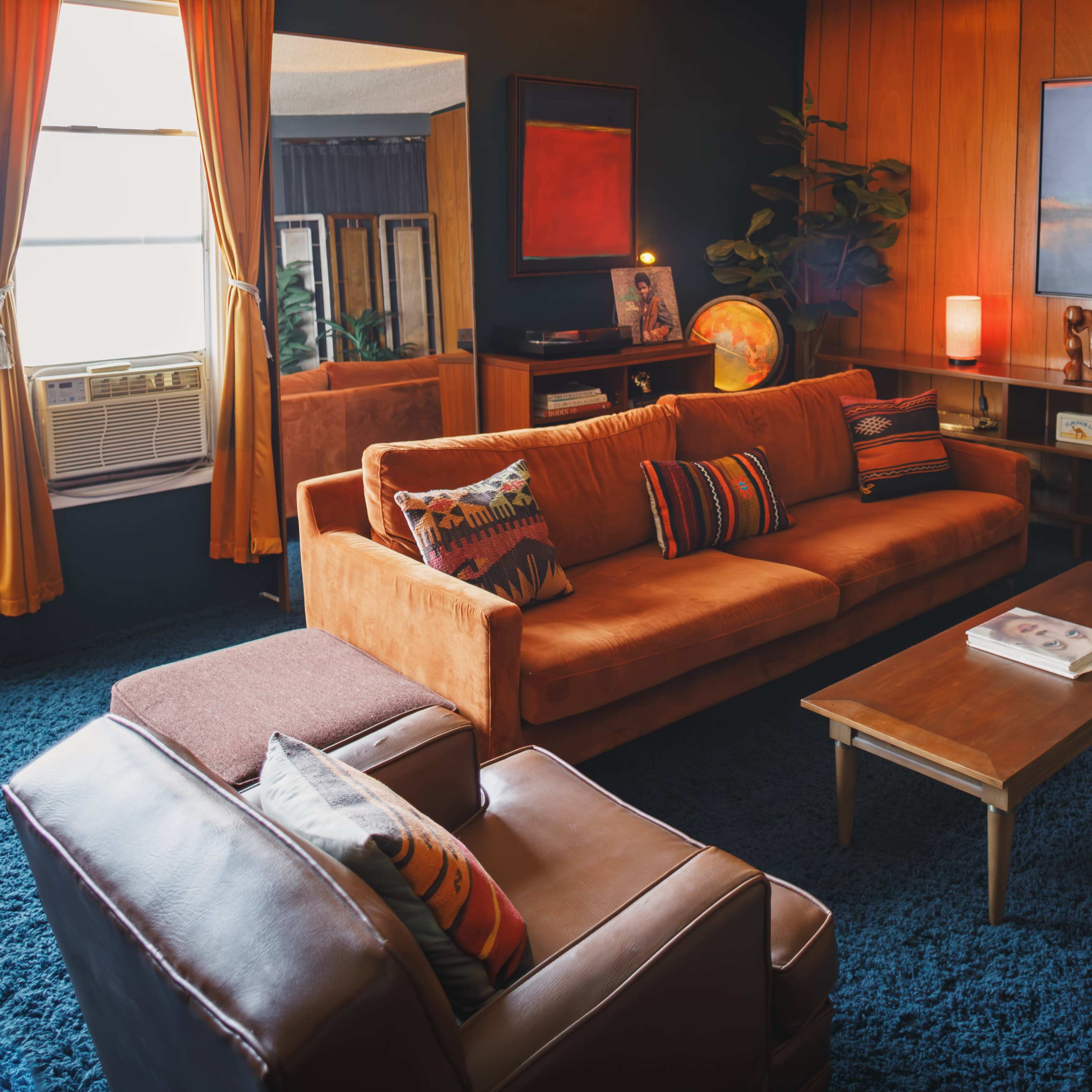 A retro-style living room featuring an orange sofa, a wooden coffee table, and plants, with a combination of warm lighting and dark walls.