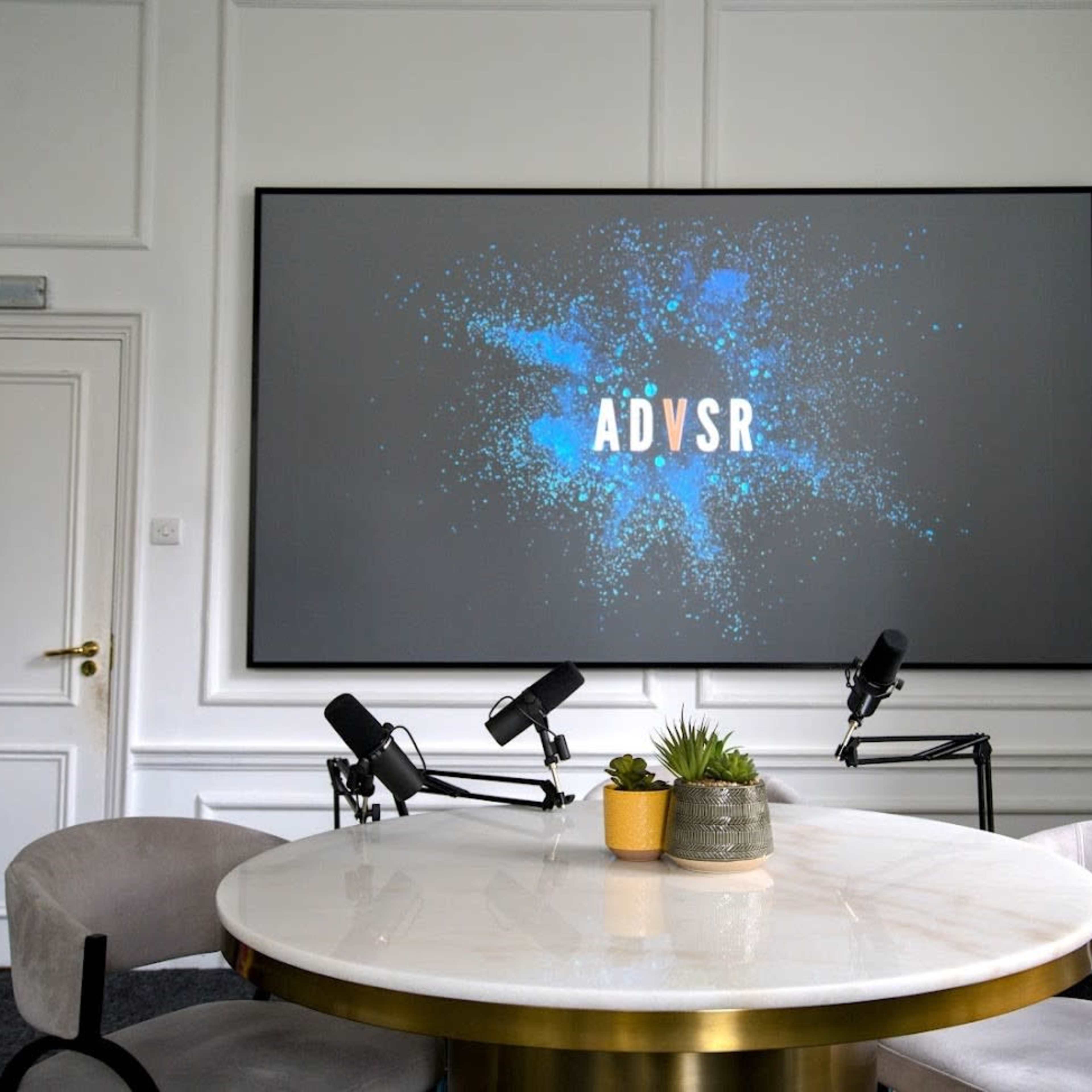 A modern meeting room features a large screen displaying "ADSVR," with two microphones and a potted plant positioned on a round table.