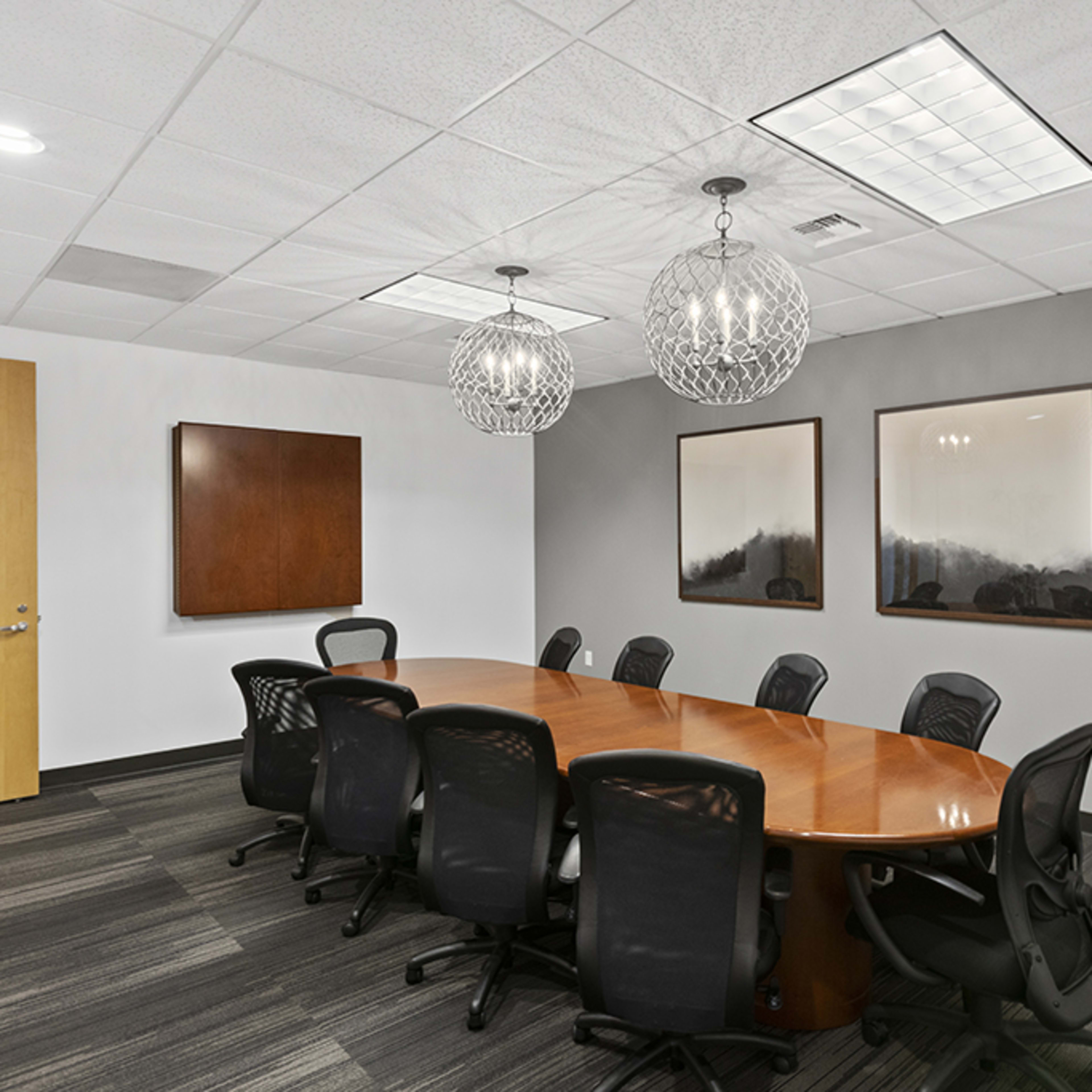 A modern conference room with a large oval wooden table surrounded by black mesh chairs, and two decorative pendant lights hanging from the ceiling.