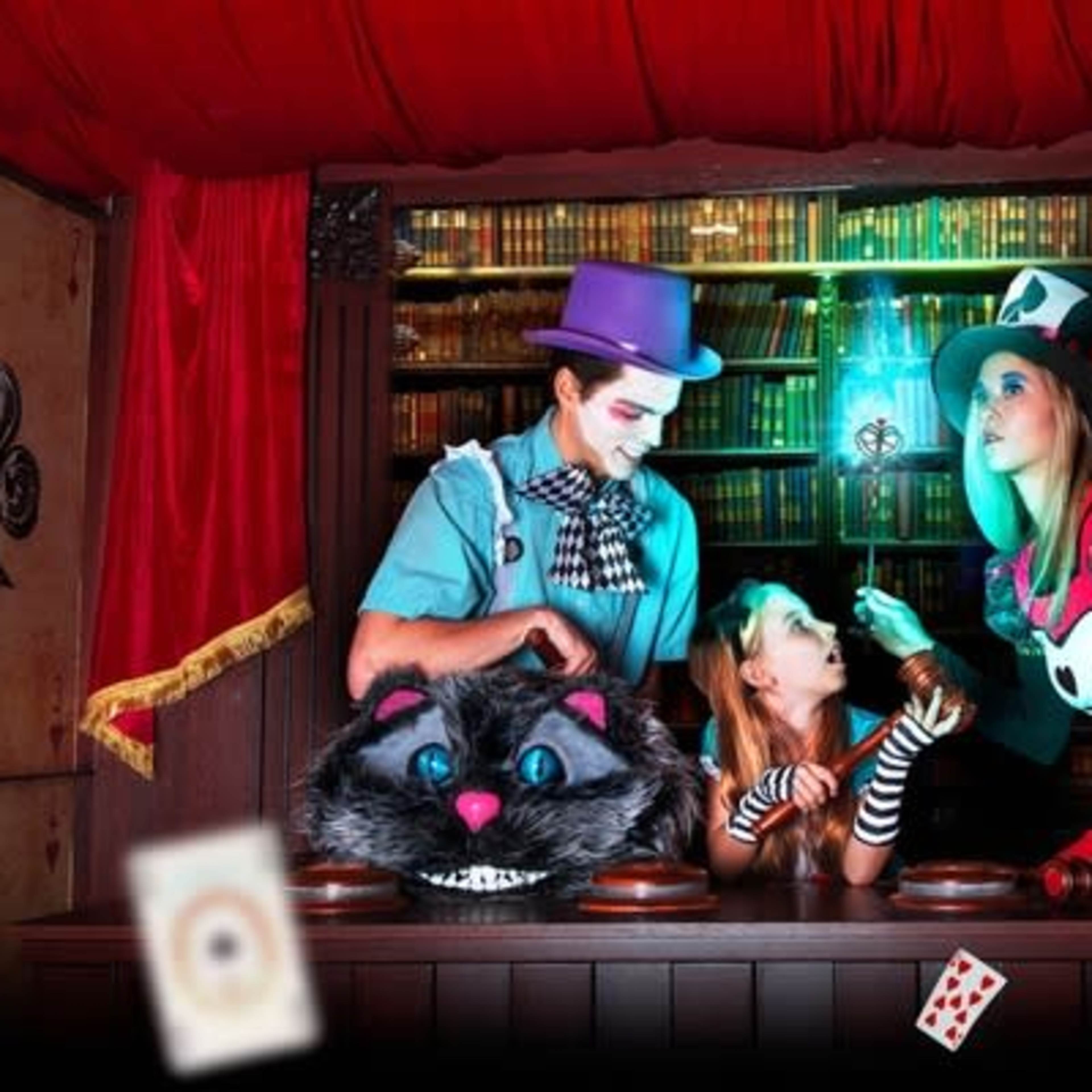 Three individuals dressed in whimsical costumes stand behind a wooden counter adorned with books and playing cards, engaged in a playful scene inspired by a fantastical theme.