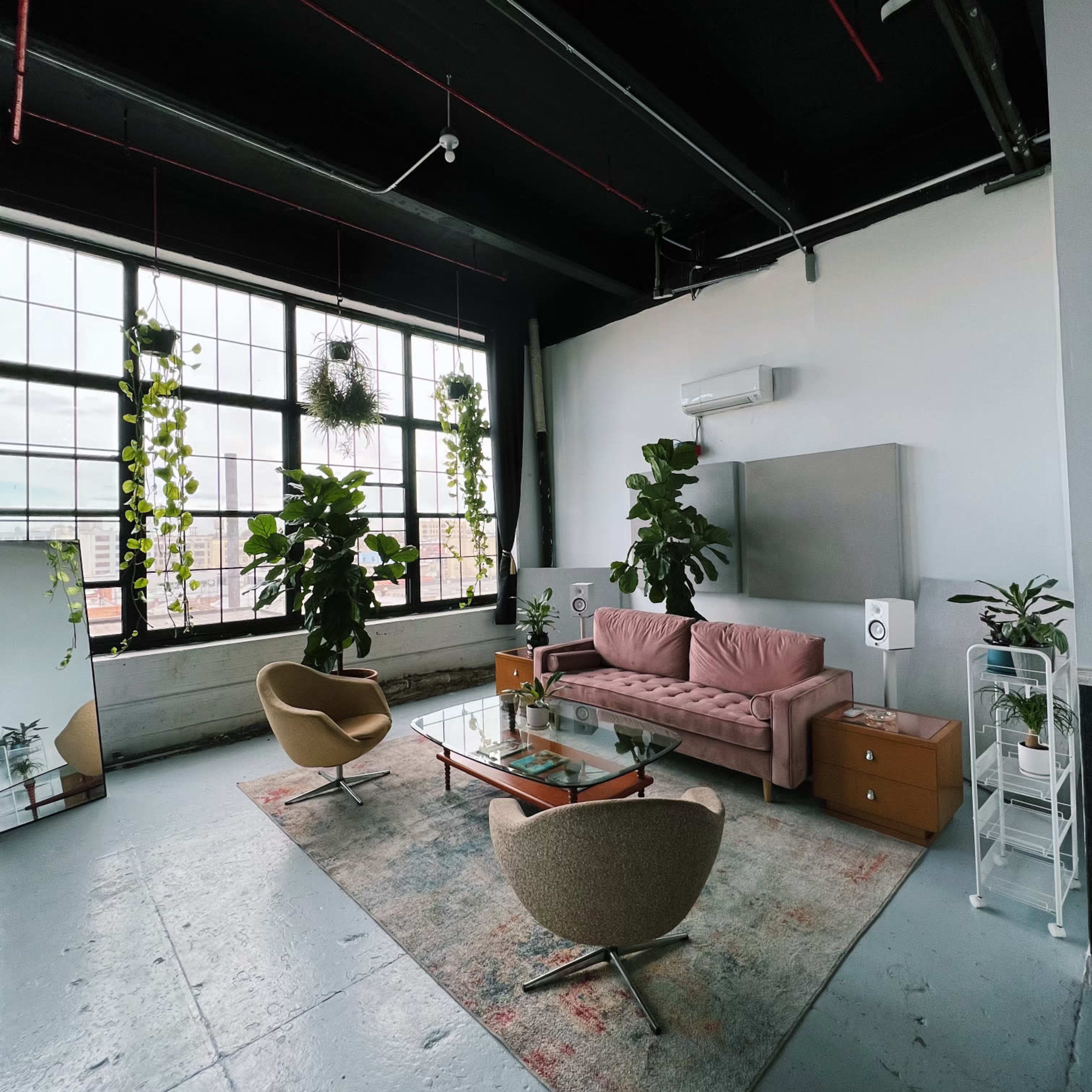 A modern living room featuring a pink sofa, two tan armchairs, indoor plants, a glass coffee table, and large windows with city views.