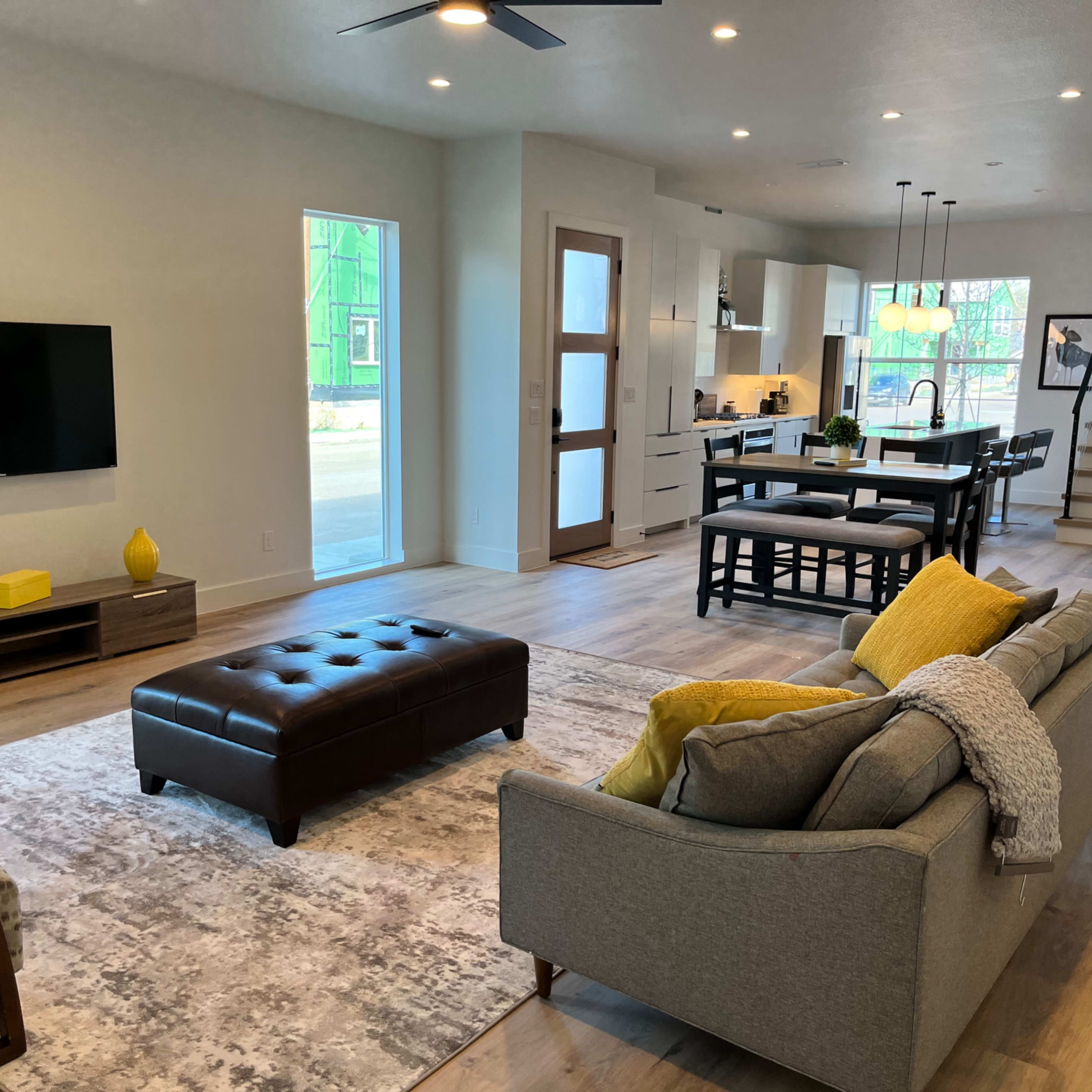 A modern living room with a flat-screen TV mounted on the wall, a dark leather ottoman in the center, and a gray sofa with yellow accents, leading to an open kitchen and dining area.