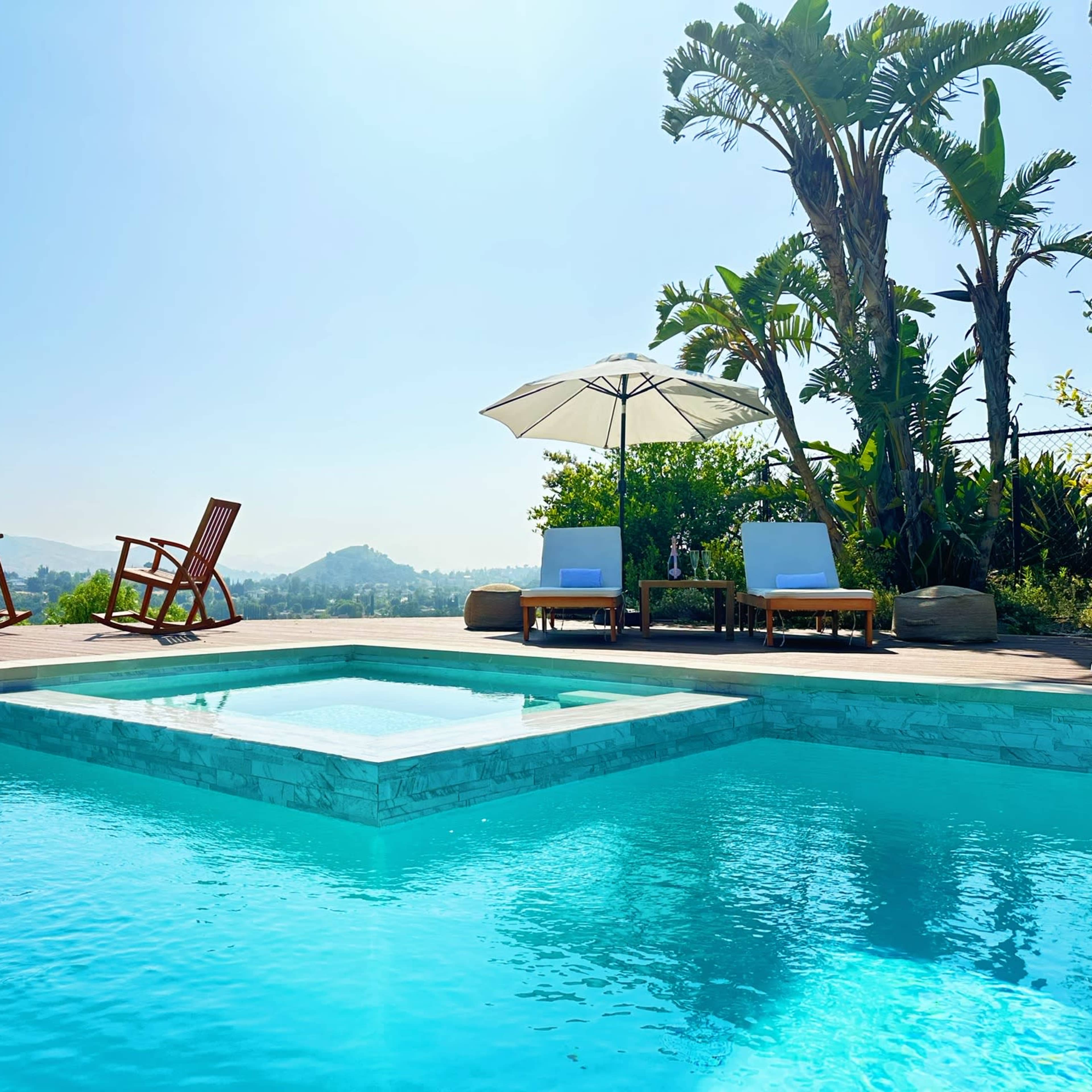The scene features a sparkling swimming pool surrounded by wooden lounge chairs and a sun umbrella, set against a backdrop of hills and clear blue skies.
