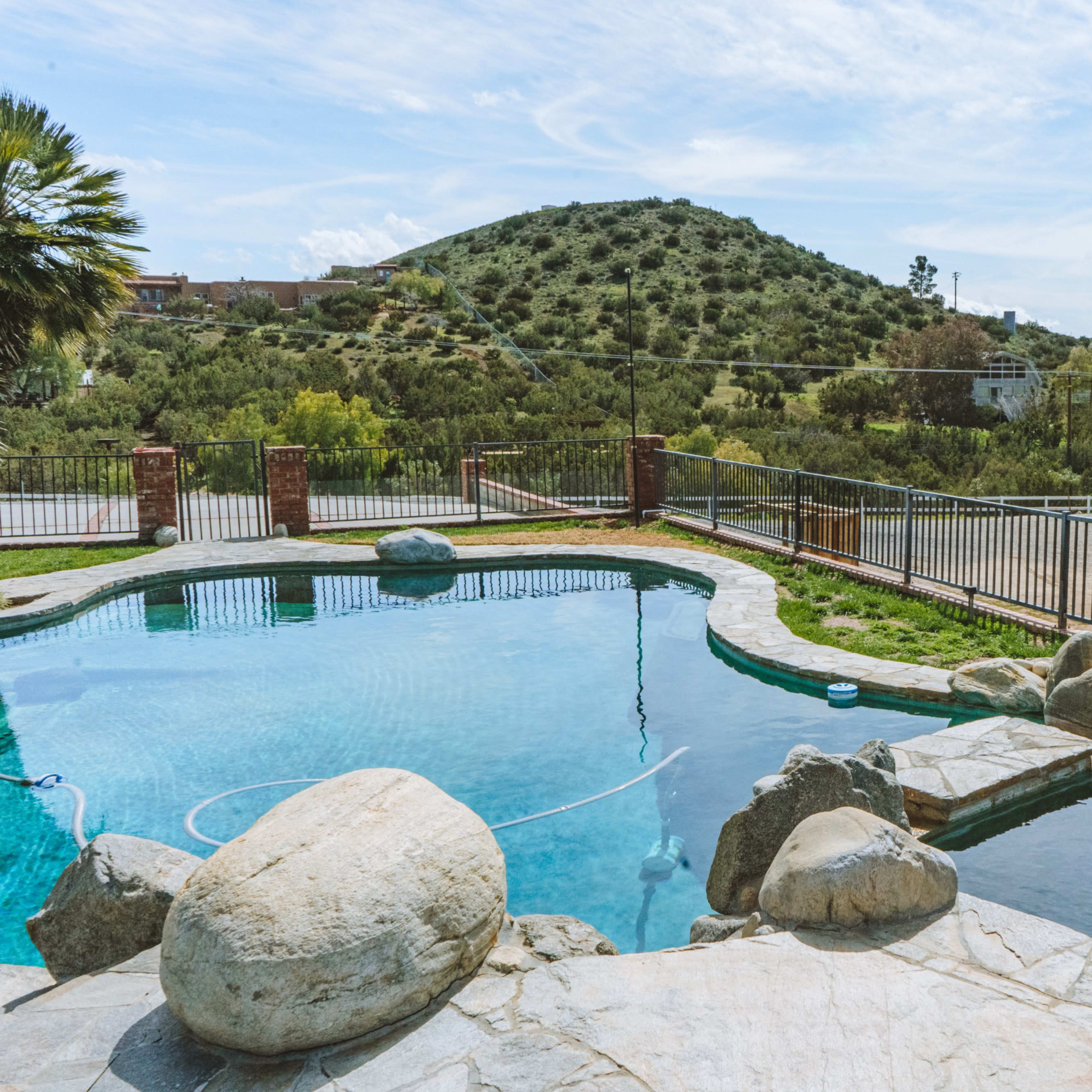 A swimming pool surrounded by rocks, with a hillside and a fence in the background.