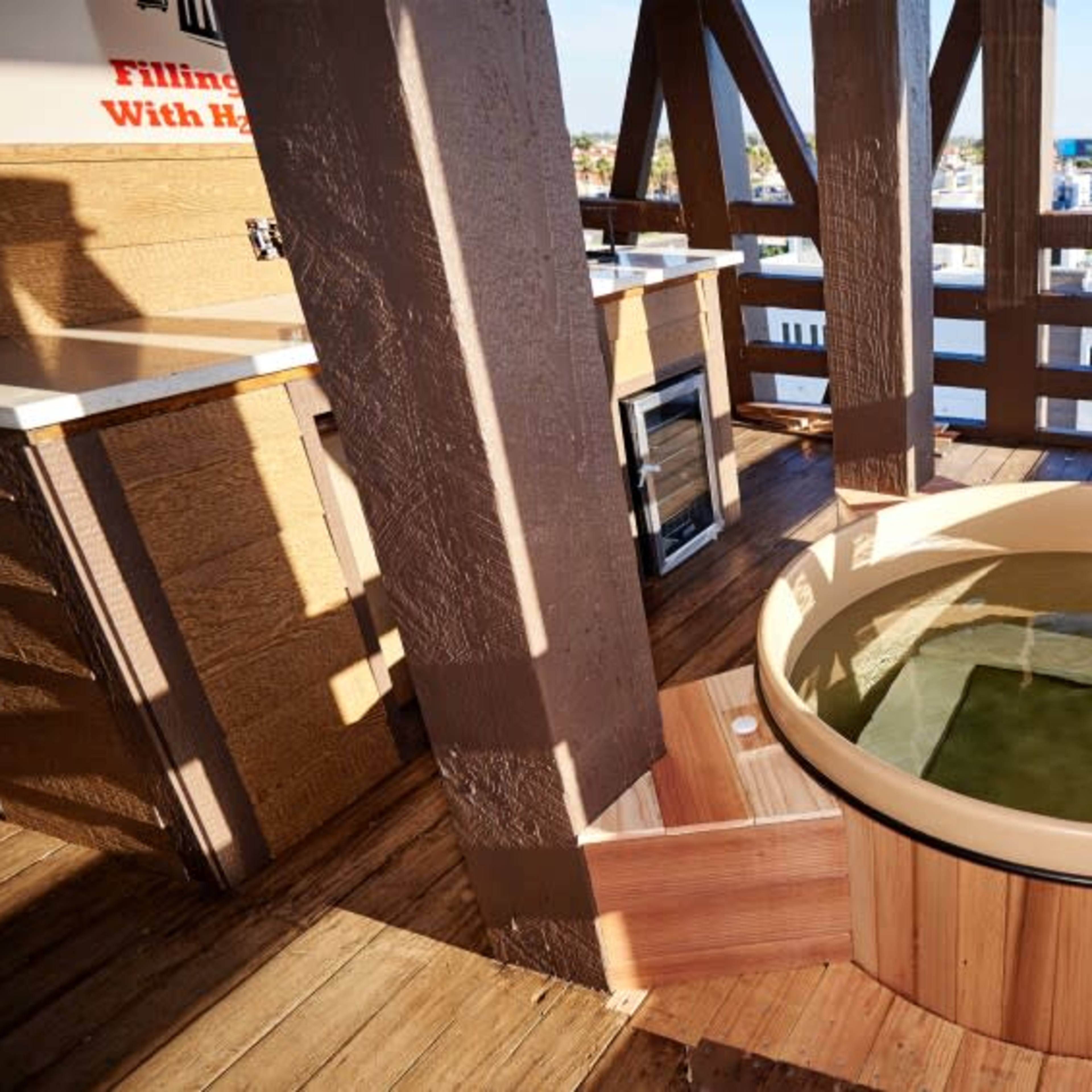 A wooden hot tub situated on a deck with a view of a coastal town and the ocean in the background.