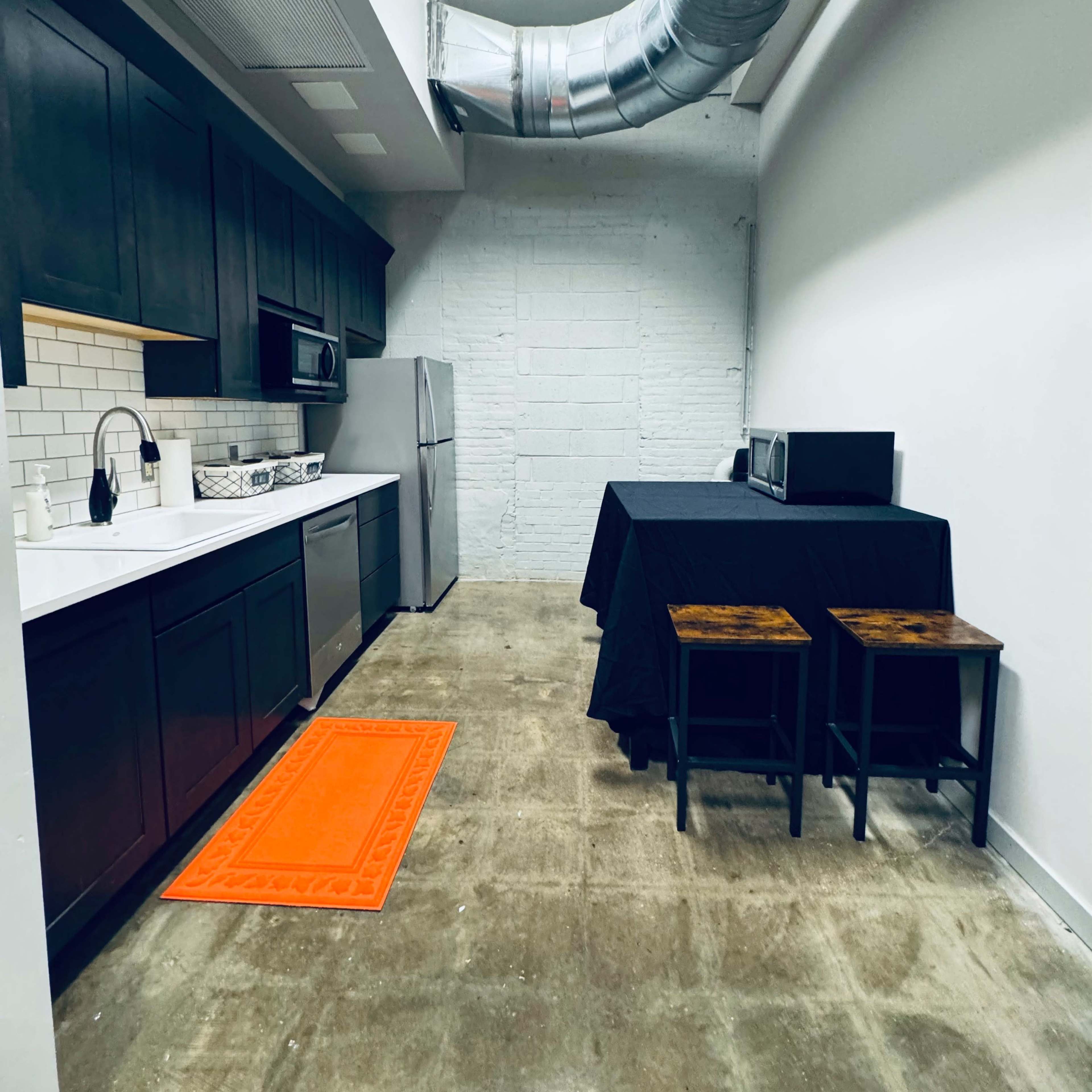 A narrow kitchen with dark cabinetry, a stainless steel refrigerator, a microwave, a black table with two stools, and an orange rug on a concrete floor.