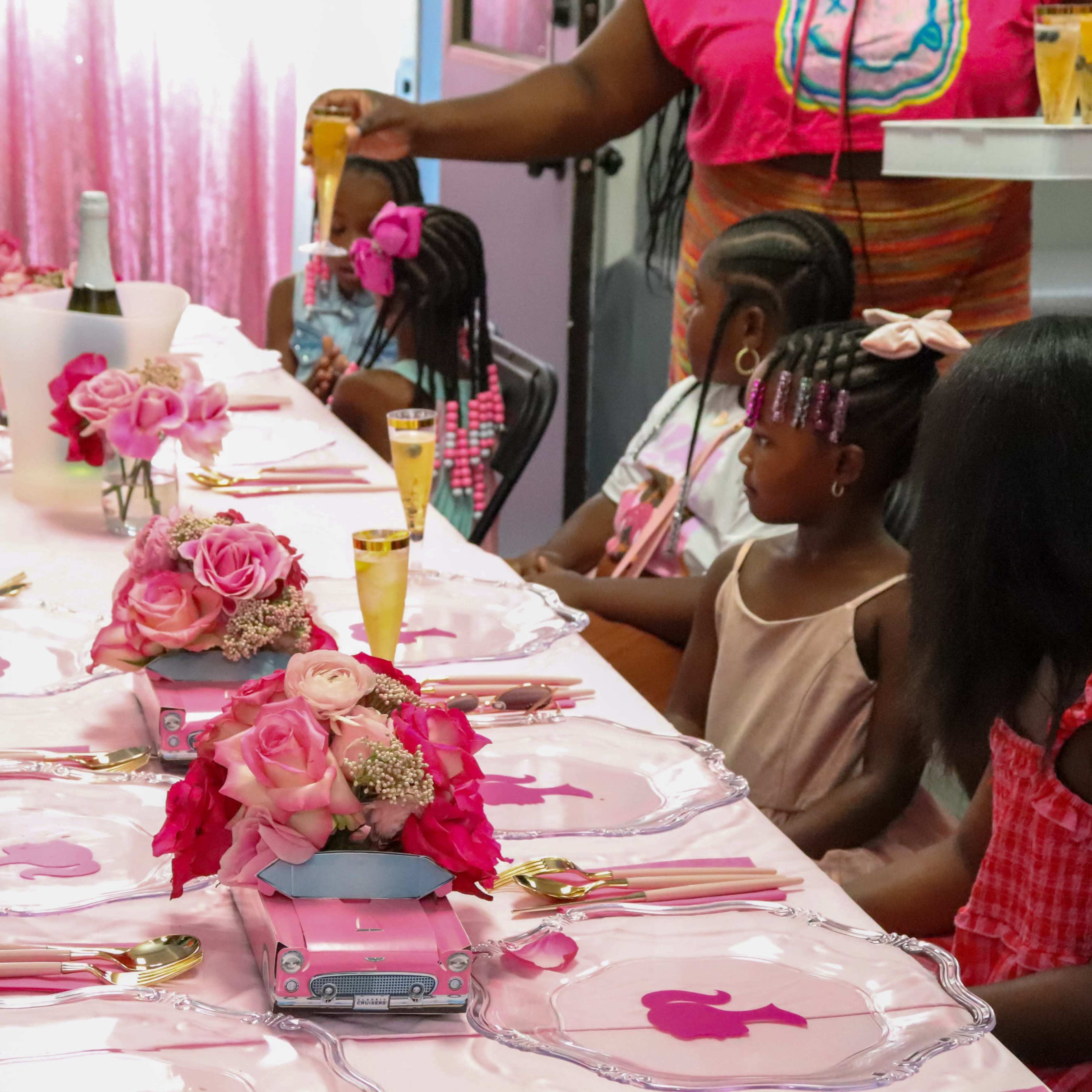 A table is set for a party with pink decorations, featuring floral centerpieces and several girls seated nearby.