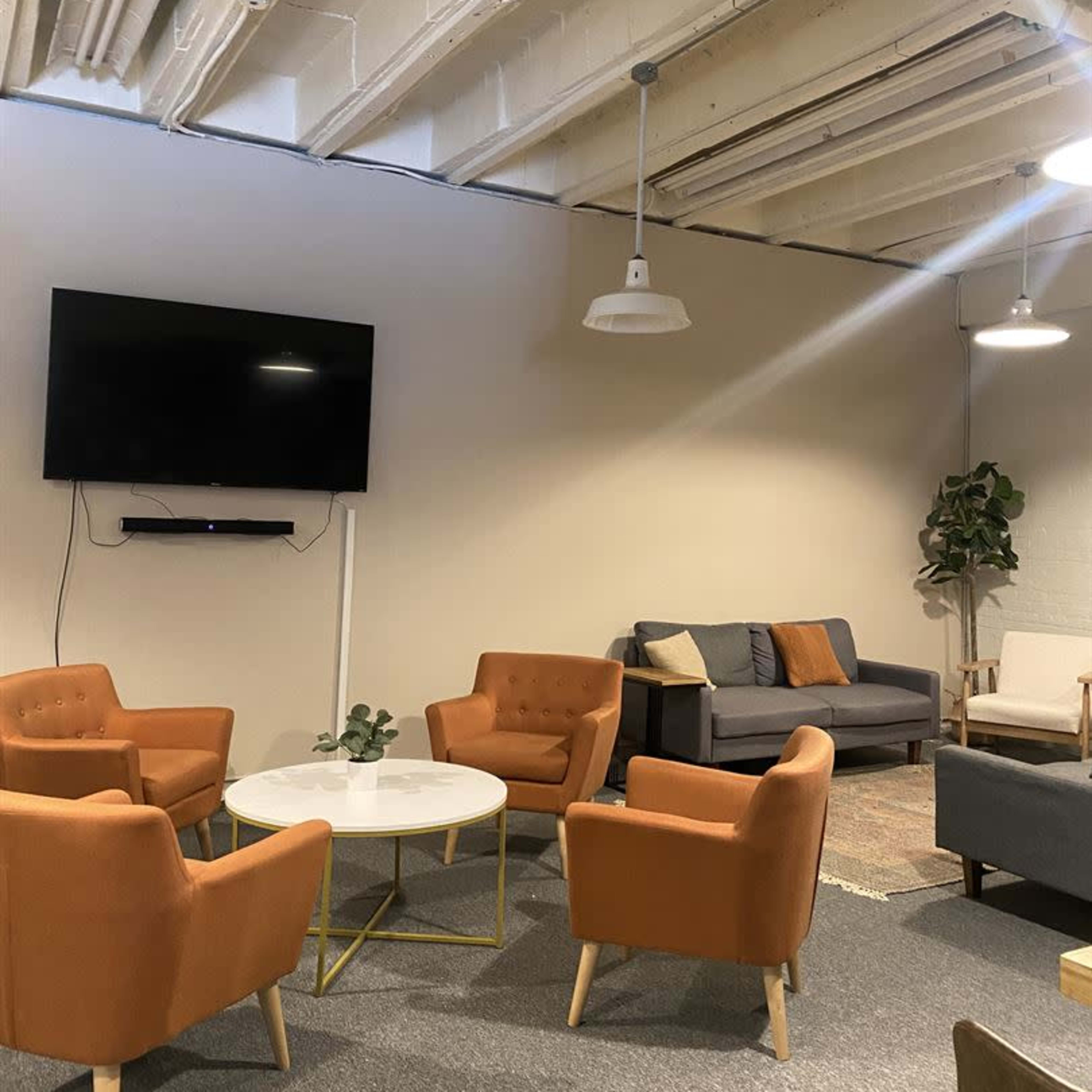 A lounge area with four orange chairs surrounding a circular coffee table, a gray couch against the wall, and a mounted television above the seating.