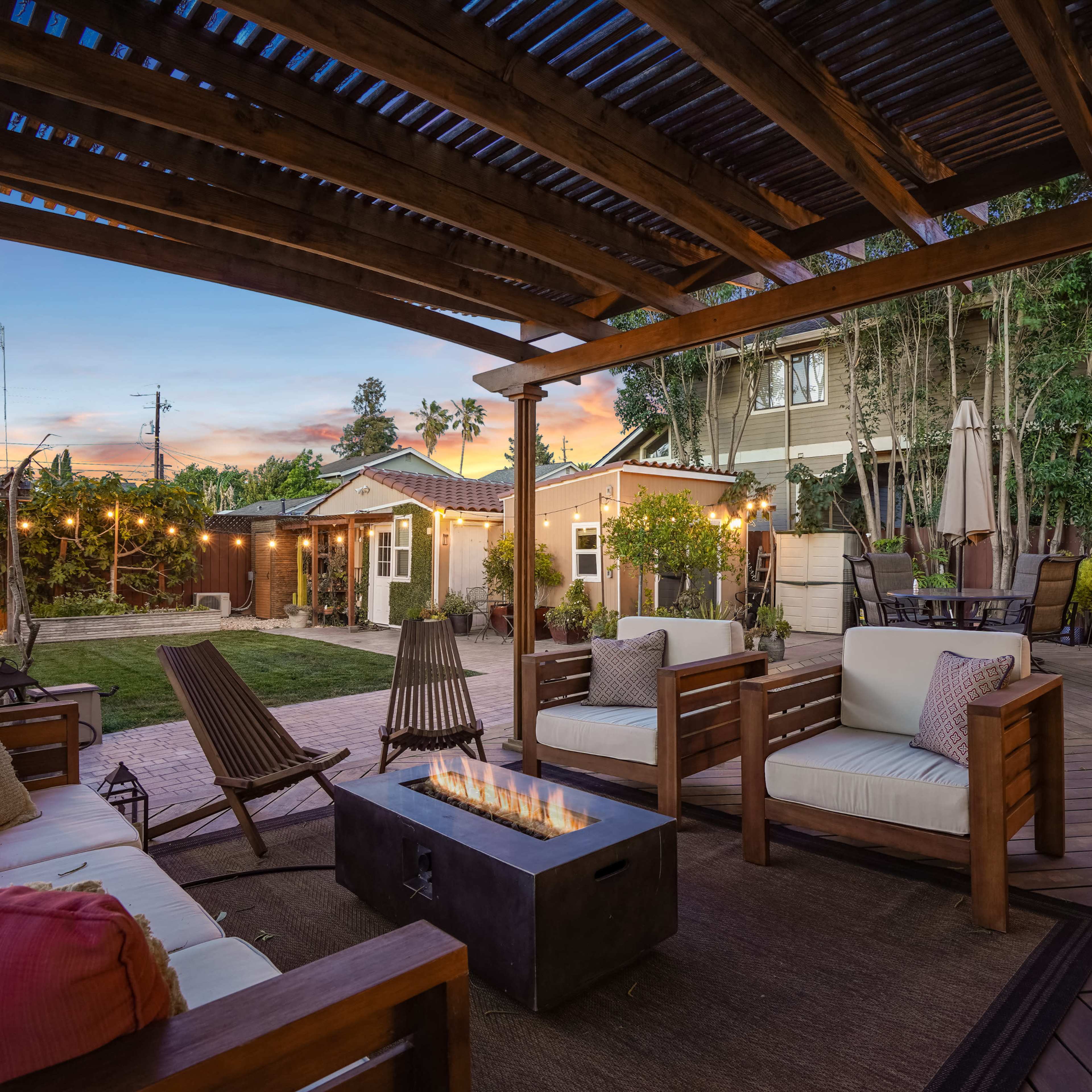 A landscaped outdoor patio features wooden seating, a fire table, and a grassy area with adjacent garden structures under a pergola.