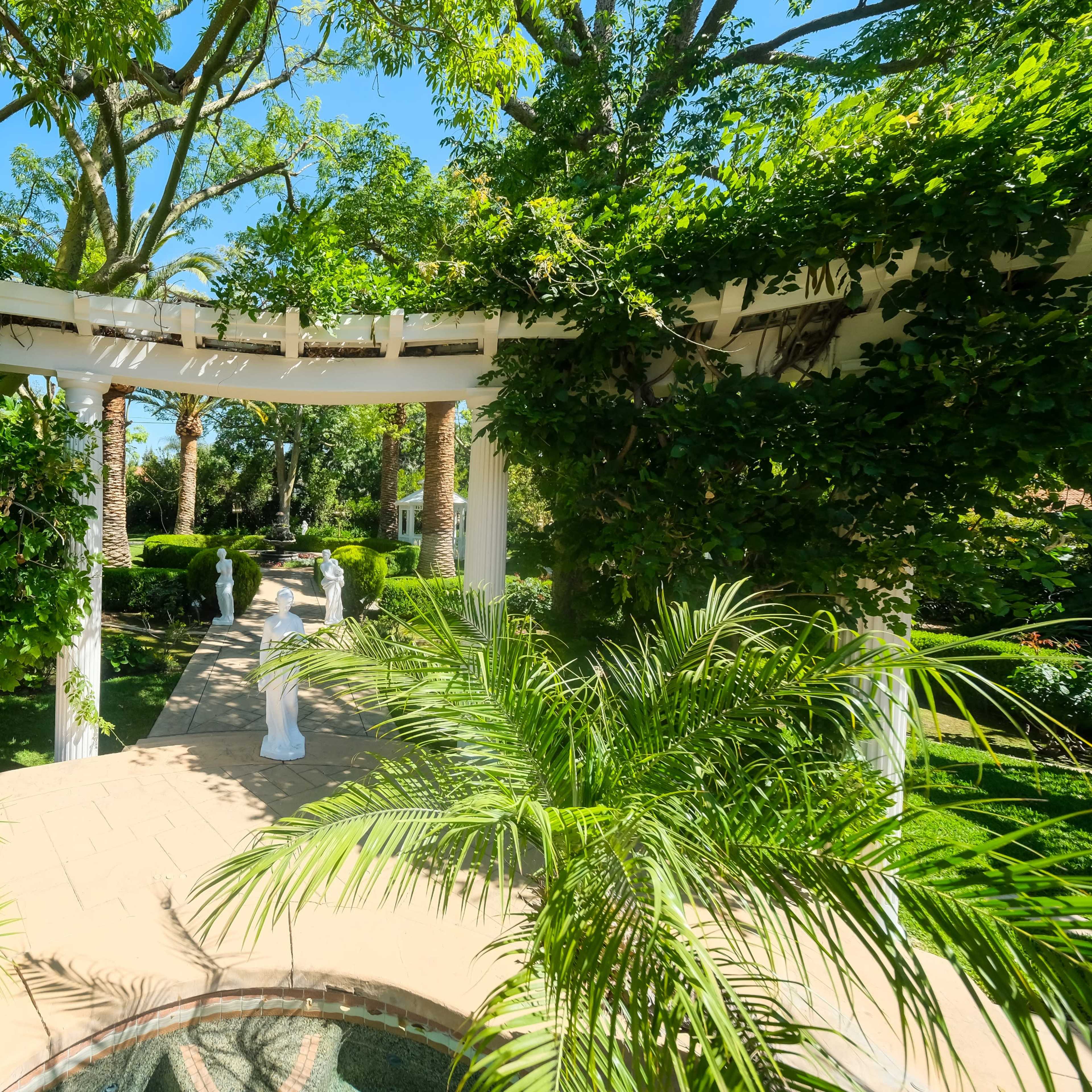 A sunlit garden features a winding path surrounded by lush greenery and decorative statues under a trellis.