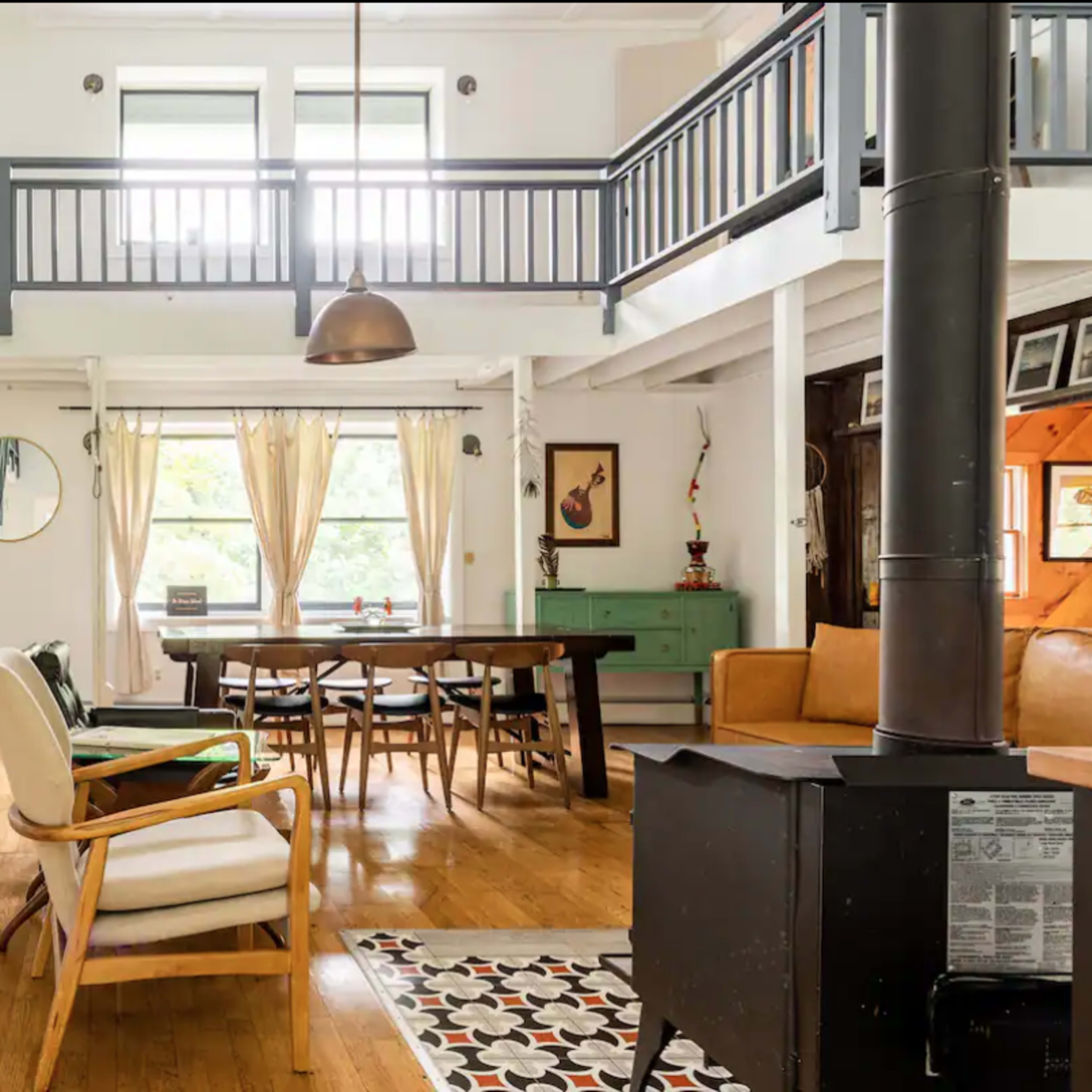 A spacious living area with wooden floors, large windows, and a dining table surrounded by chairs, complemented by a staircase leading to a lofted space above.