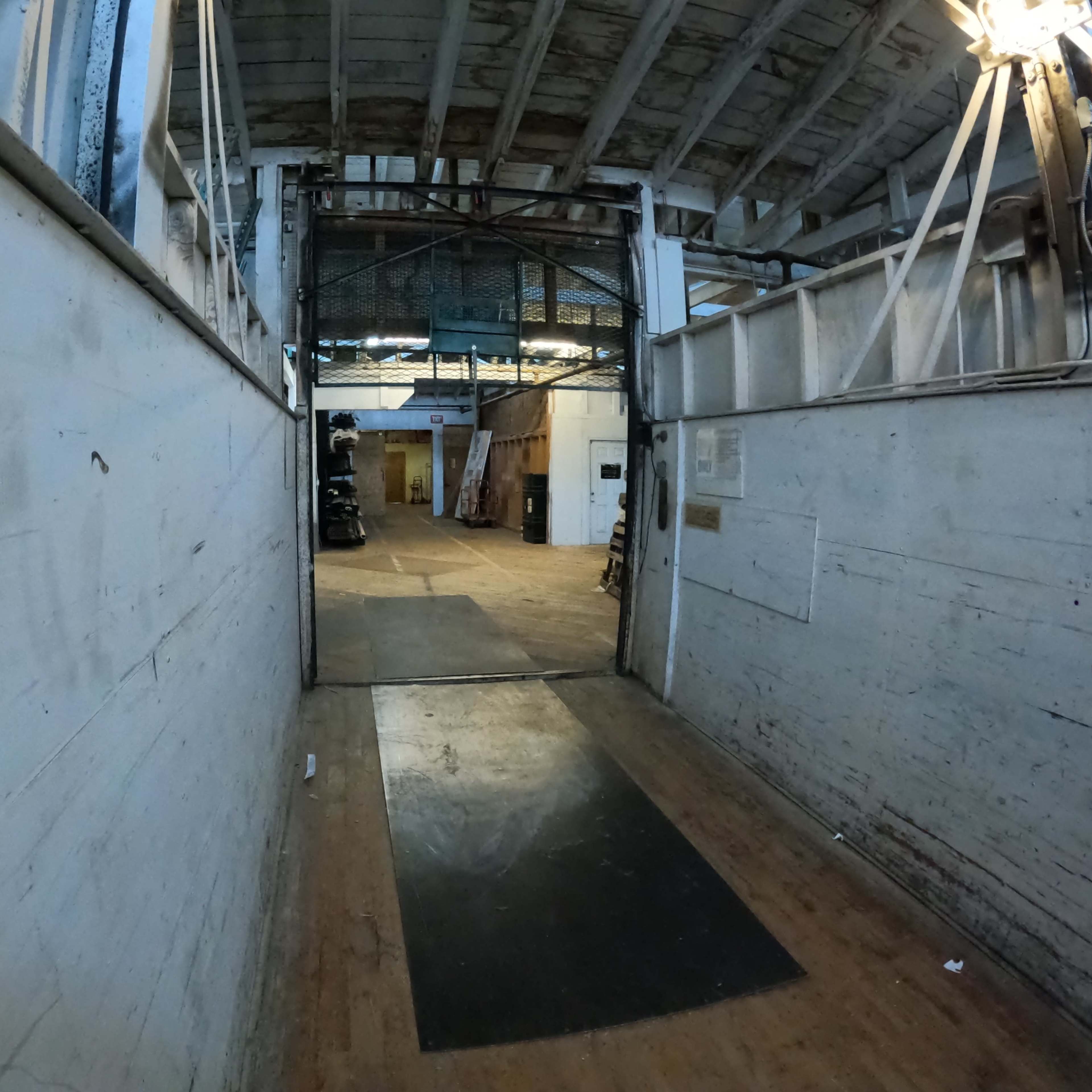 A dimly lit hallway leading to an open area with a wooden floor and various items stacked in the background.