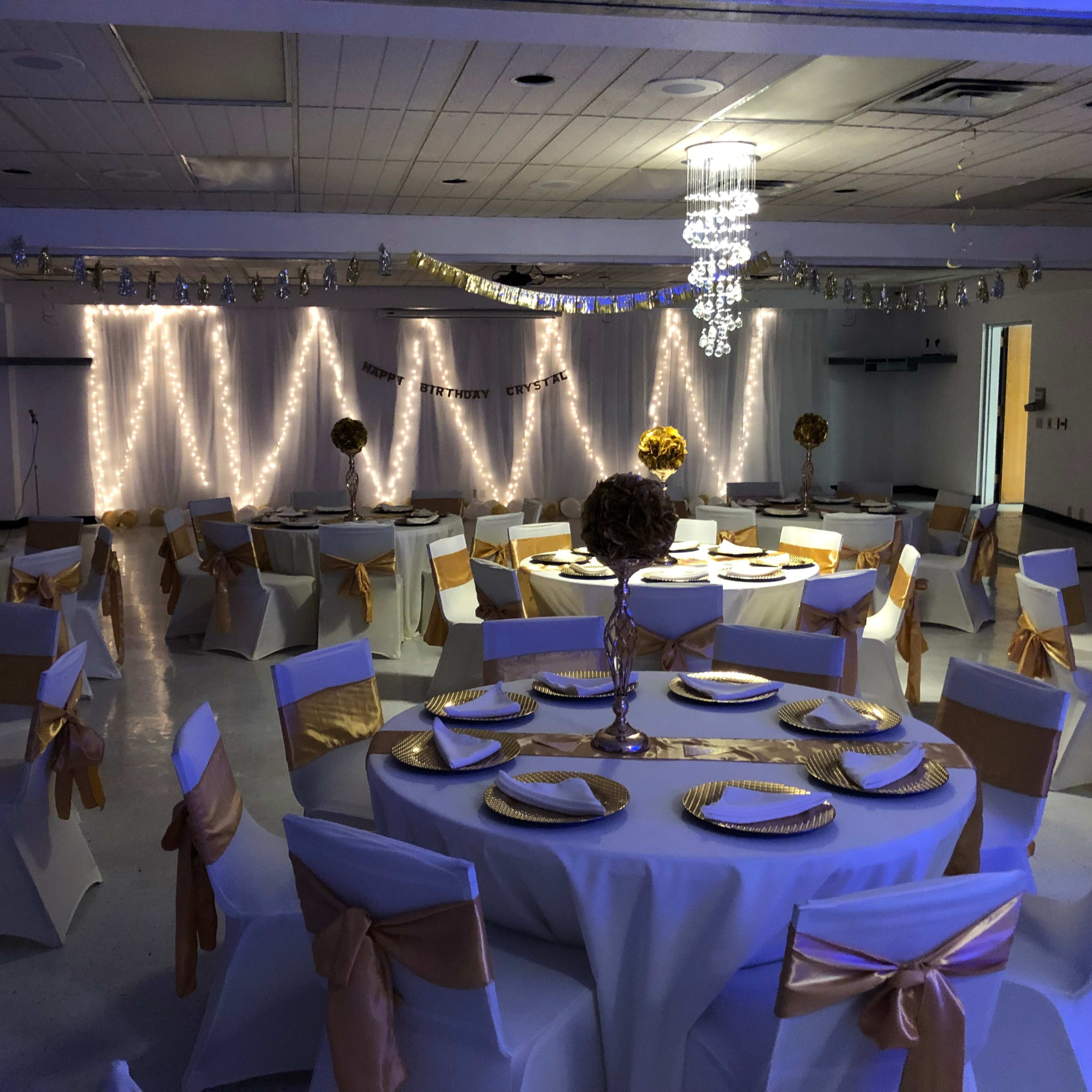 A decorated hall set up for an event, featuring round tables with white tablecloths and gold accents, surrounded by string lights and ornamental centerpieces.