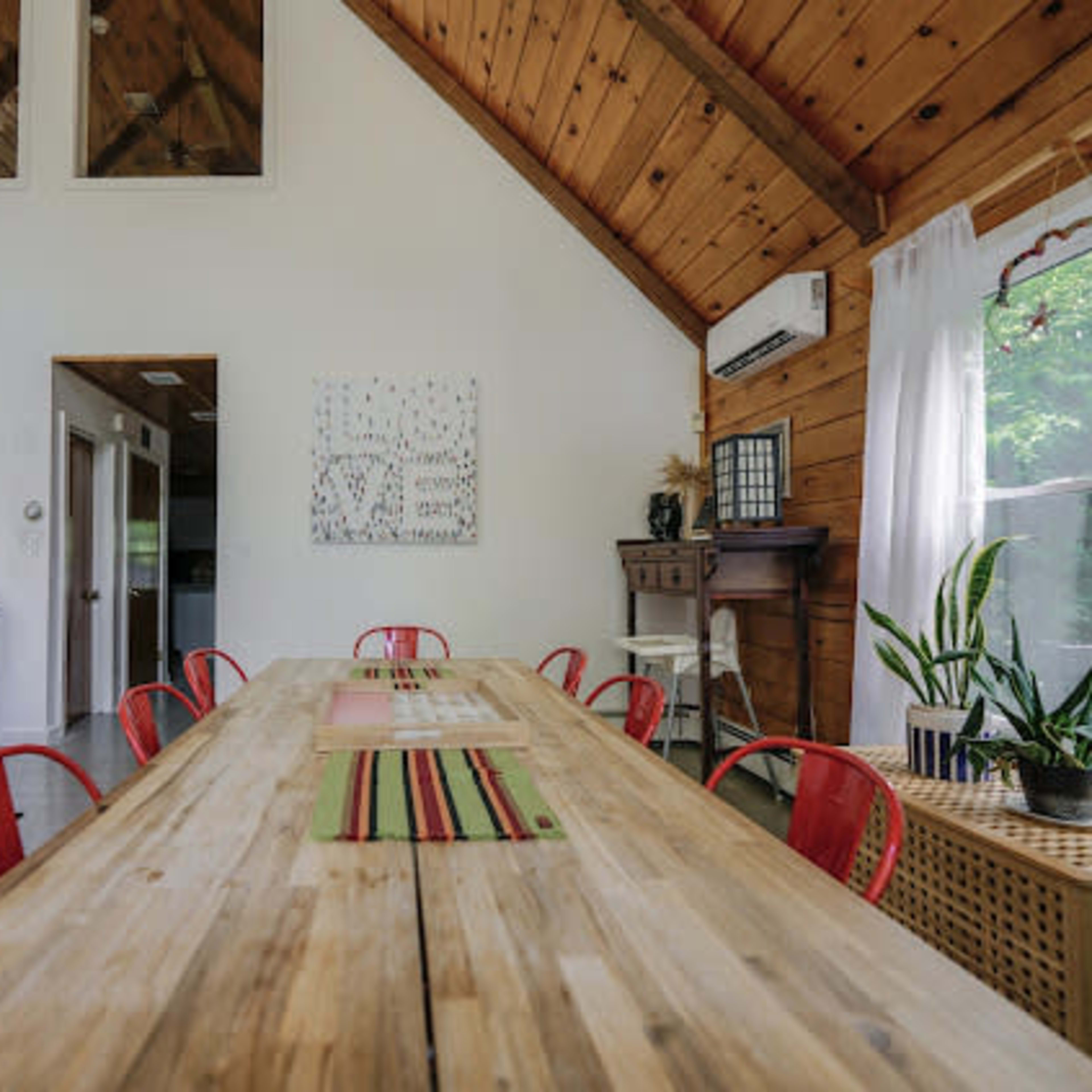 A spacious dining area features a long wooden table surrounded by red chairs, with a cozy couch and large windows in the background.