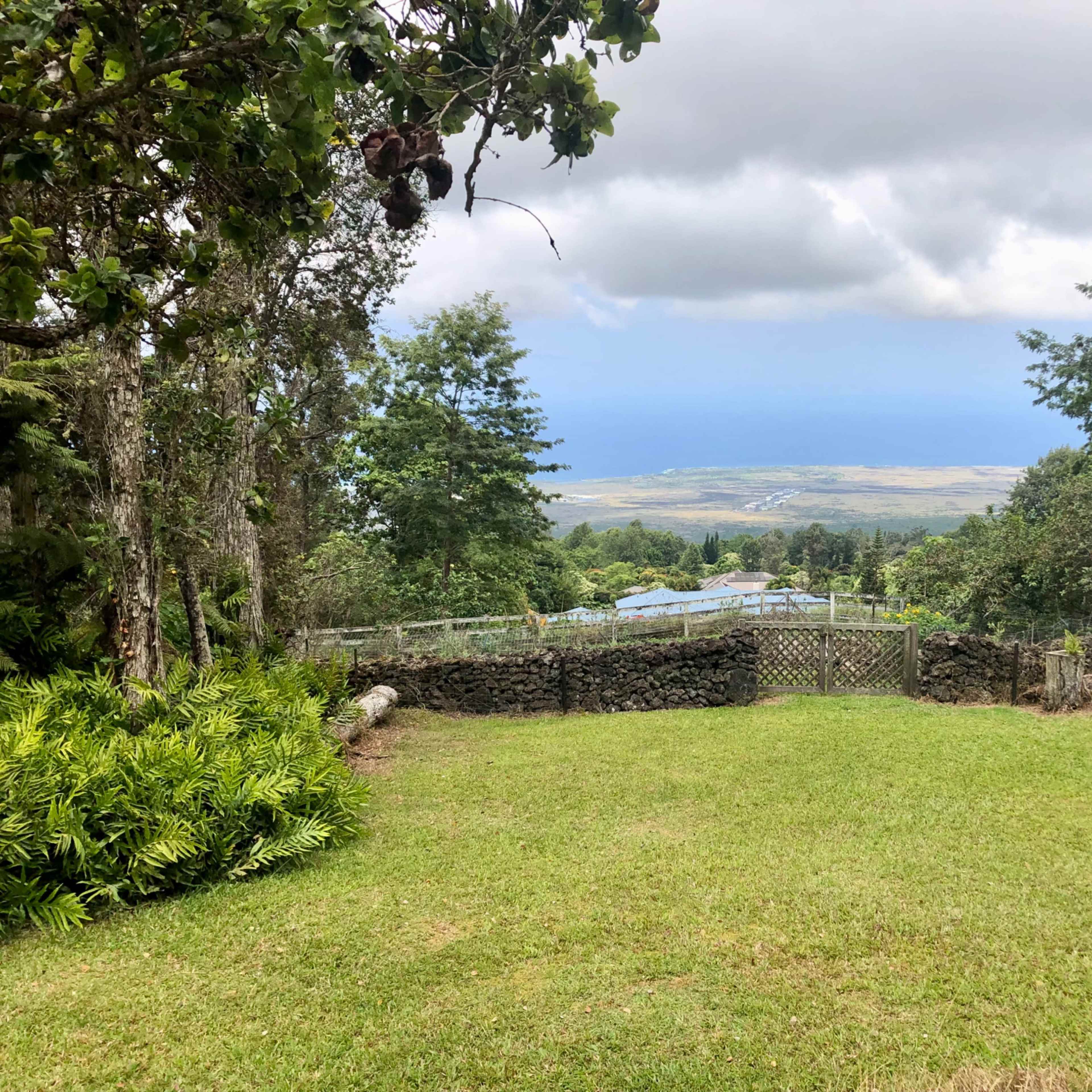 A grassy area bordered by trees, overlooking a landscape with distant hills under a cloudy sky.