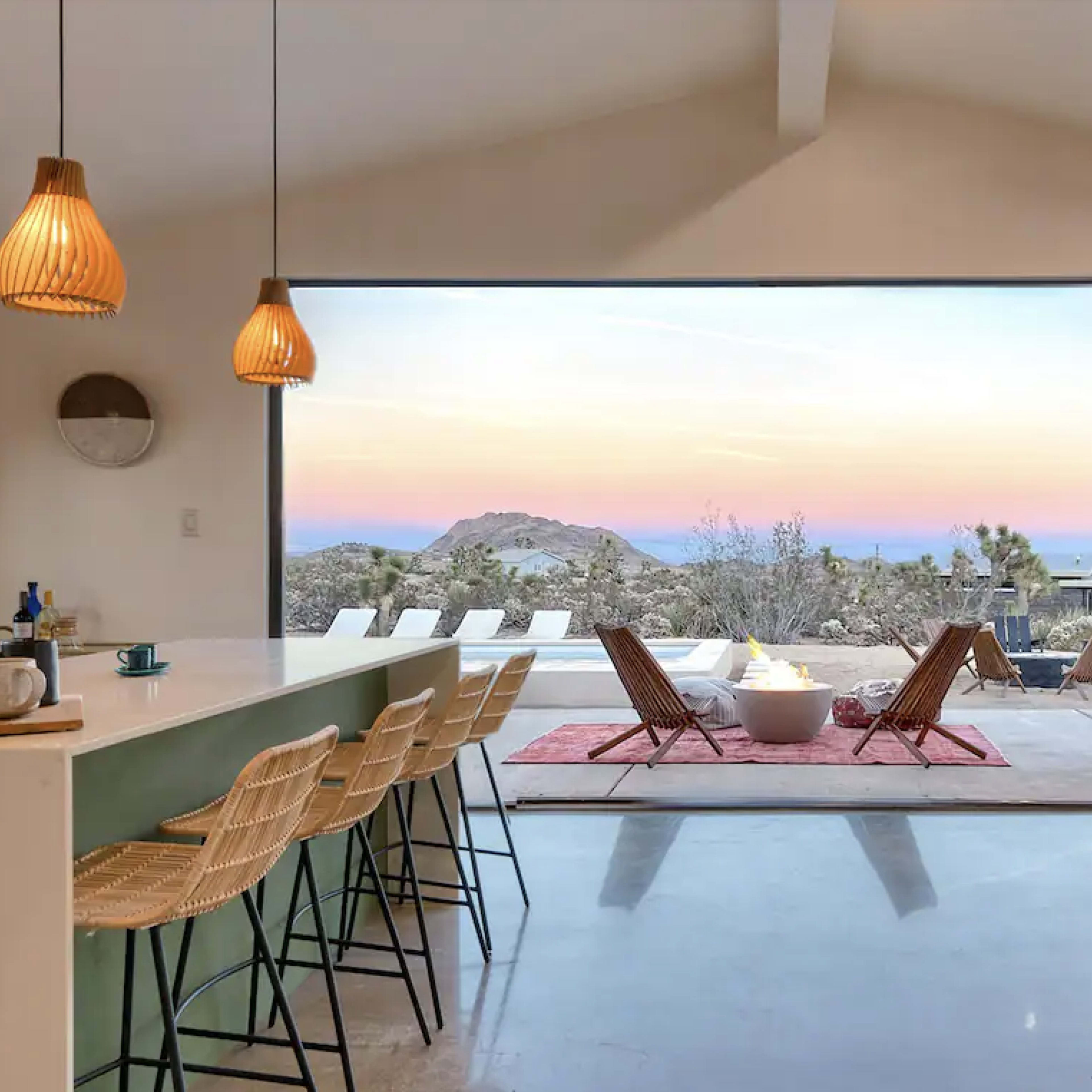 A modern interior kitchen opens to a patio with outdoor seating and a view of a mountain at sunset.