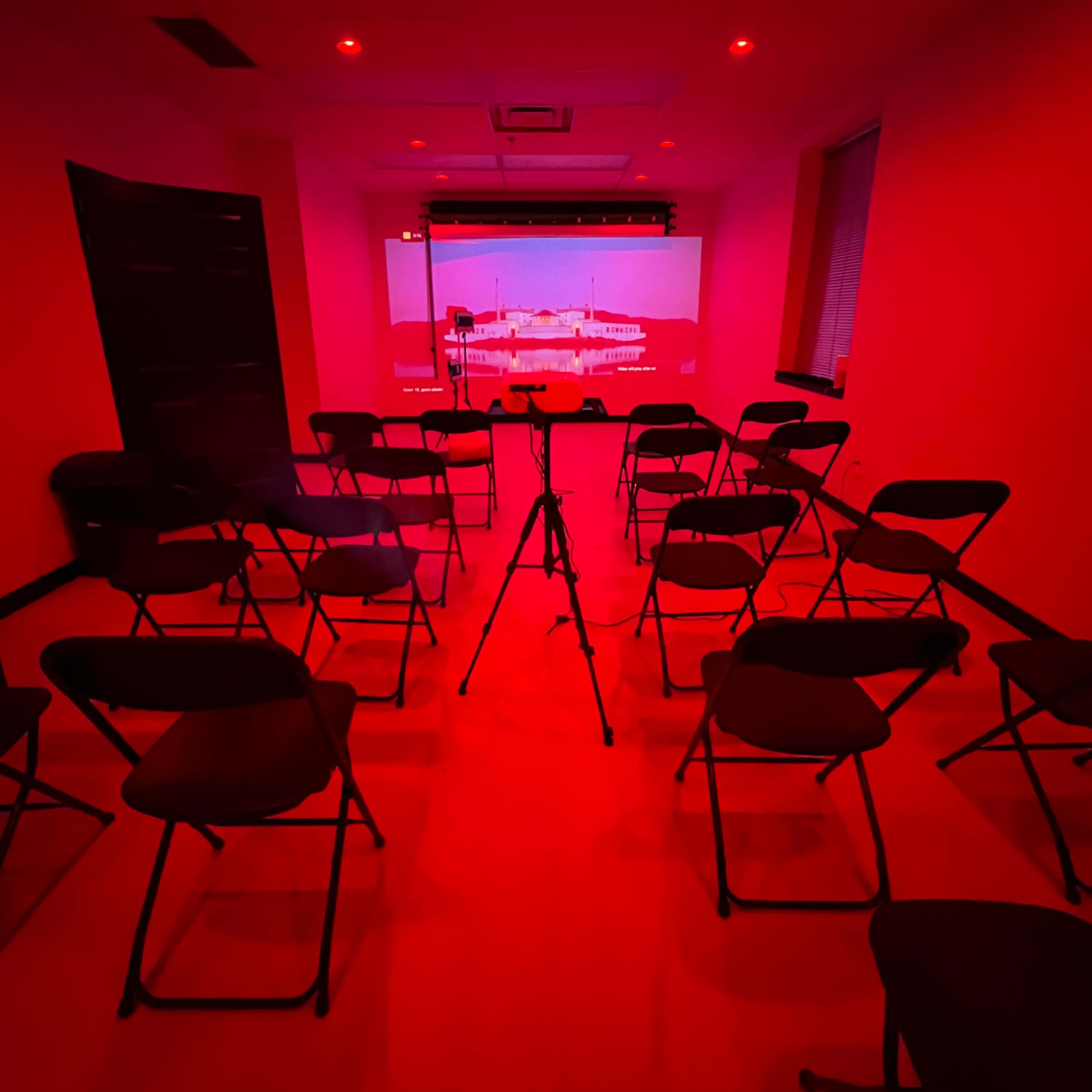 A presentation room with red lighting features arranged chairs in a circular formation around a projector screen displaying a red-tinted image.