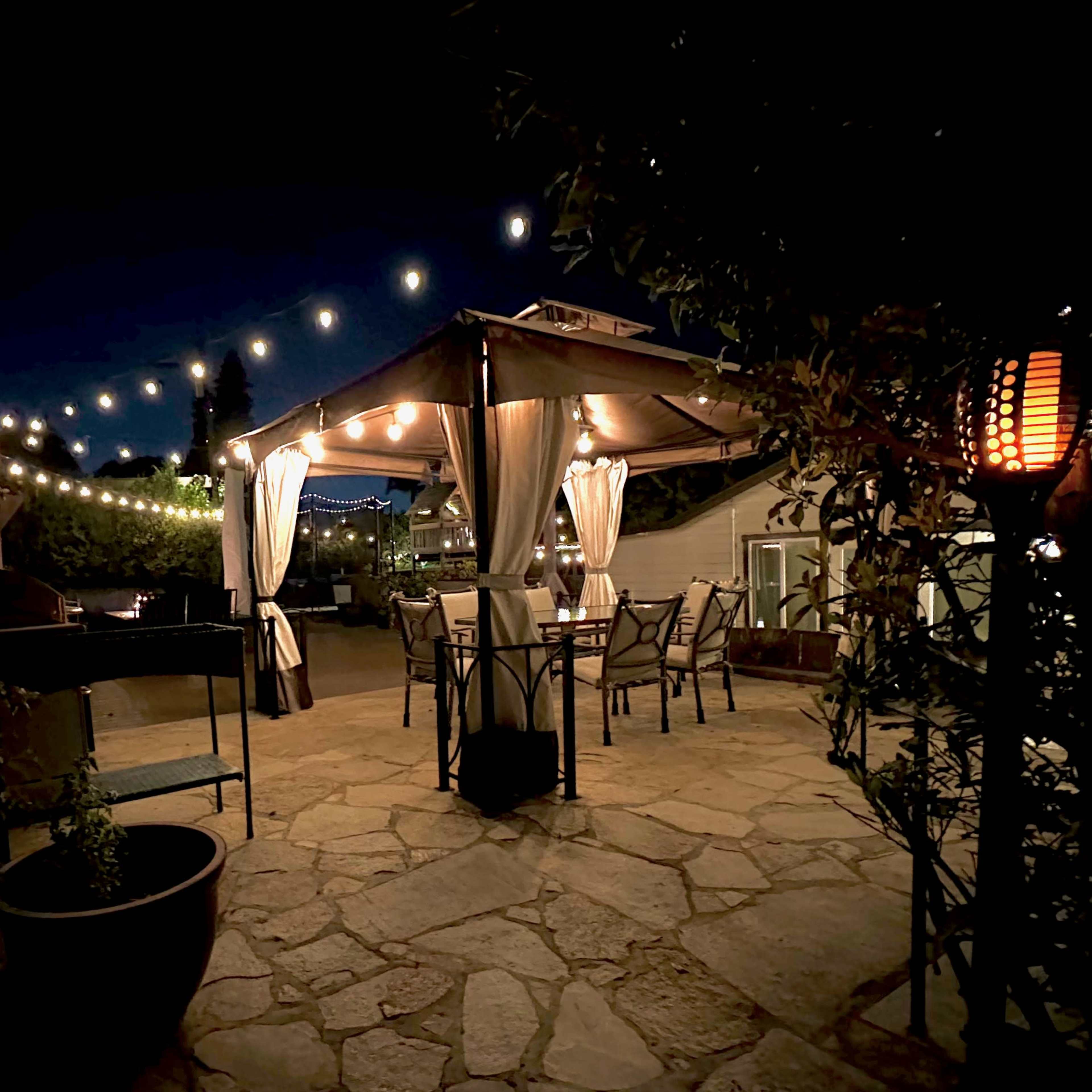 A stone-paved outdoor dining area is illuminated by string lights and lanterns, featuring a canopy over tables and chairs.