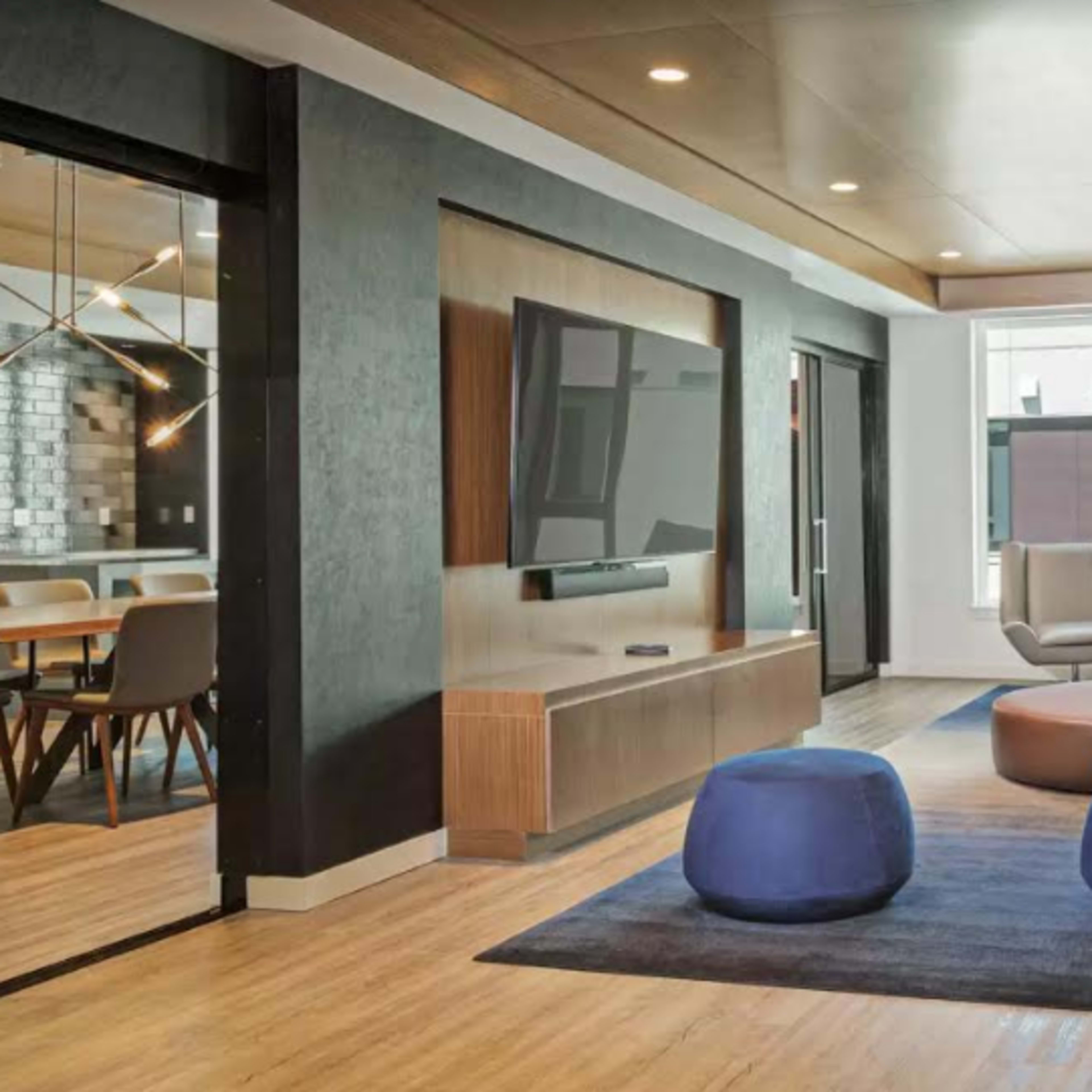 A modern lounge area with a gray sofa, two blue poufs, a wooden coffee table, and a television mounted on the wall, surrounded by large windows.