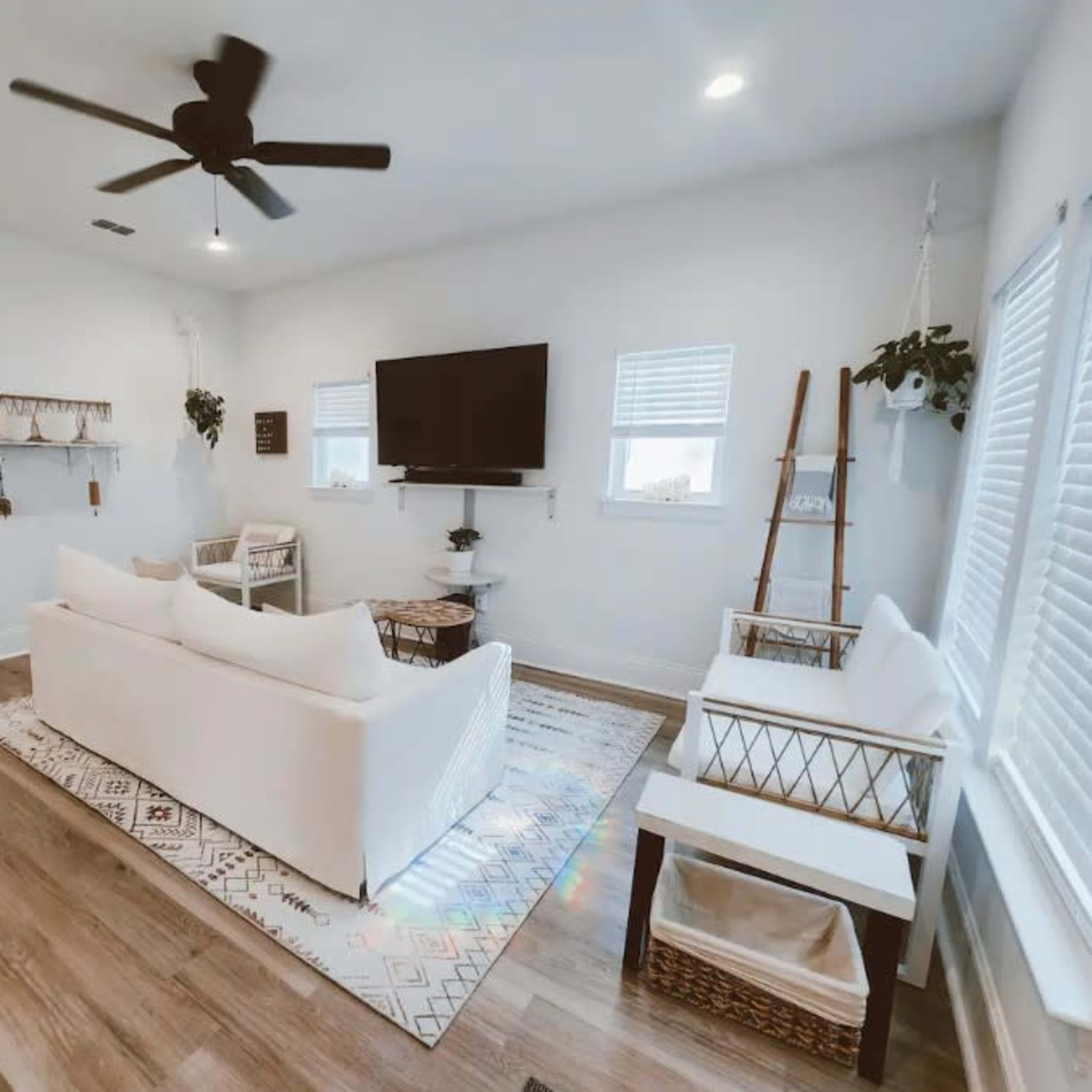 The living room features a white sofa, two chairs, a television mounted on the wall, and decorative plants arranged on shelves.