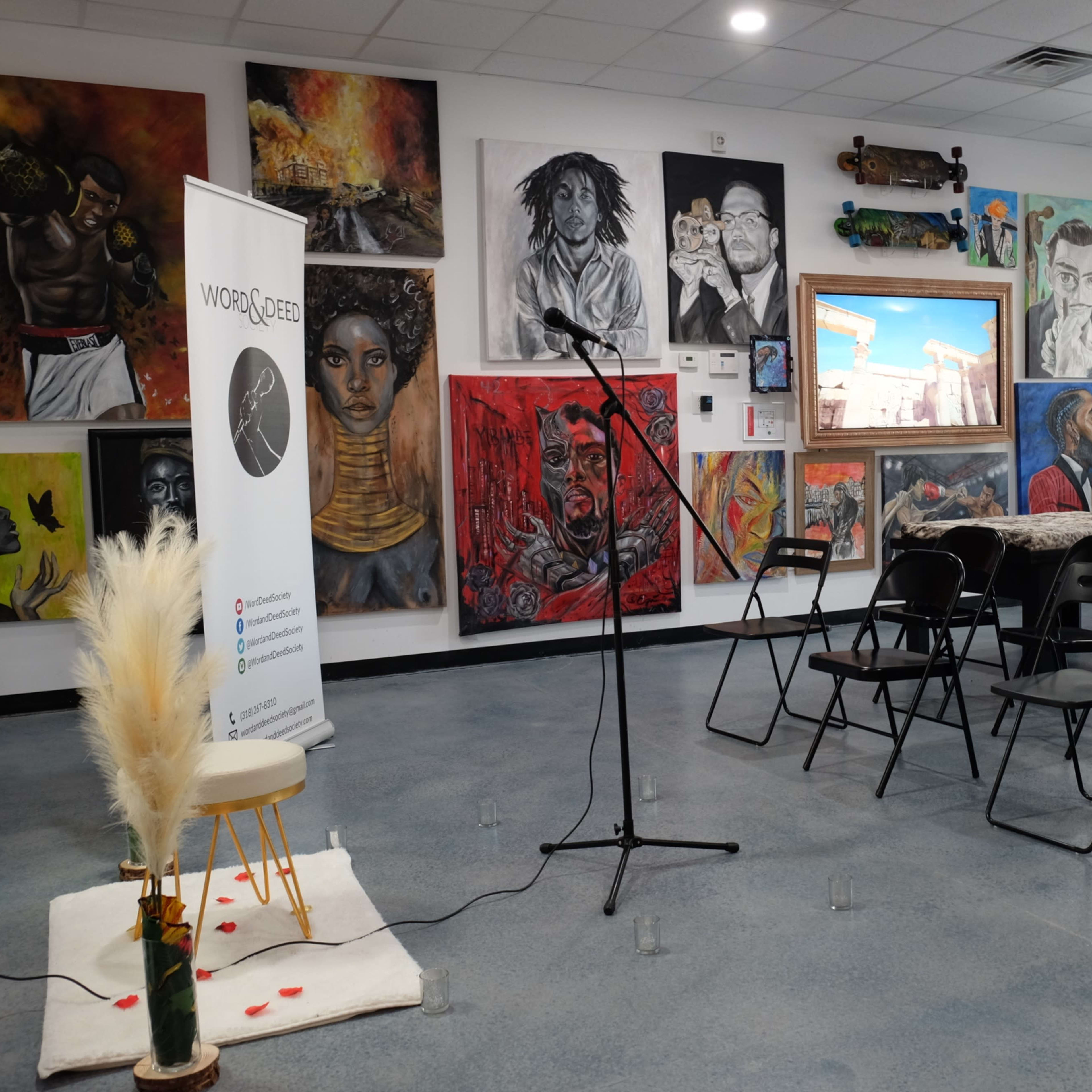 A room set up for an event, featuring empty chairs, a microphone stand, and various colorful artworks on the walls.