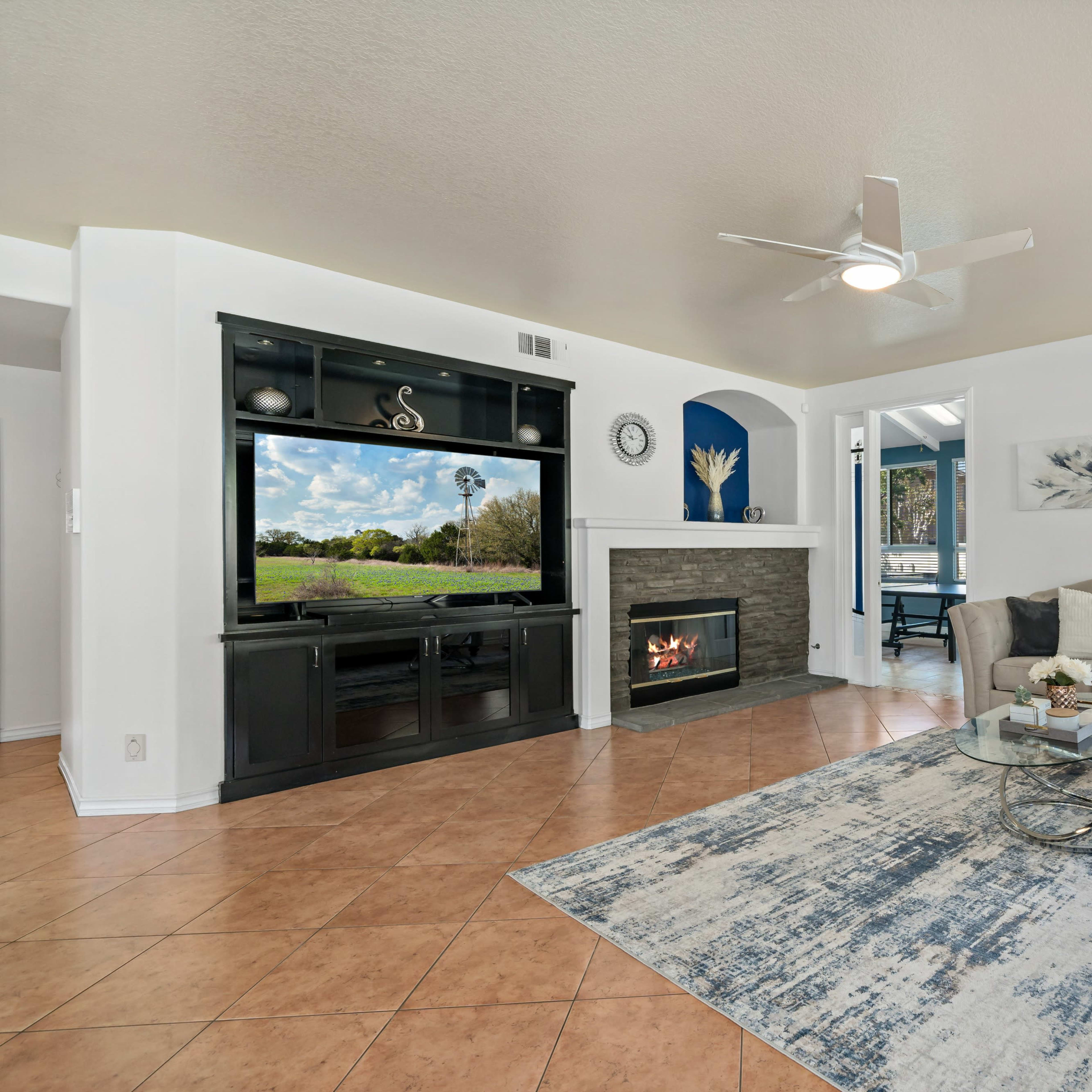 A living room features a large television above a fireplace, a light-colored sofa, a round coffee table, and a textured rug, with a view of the outdoors visible through a window.
