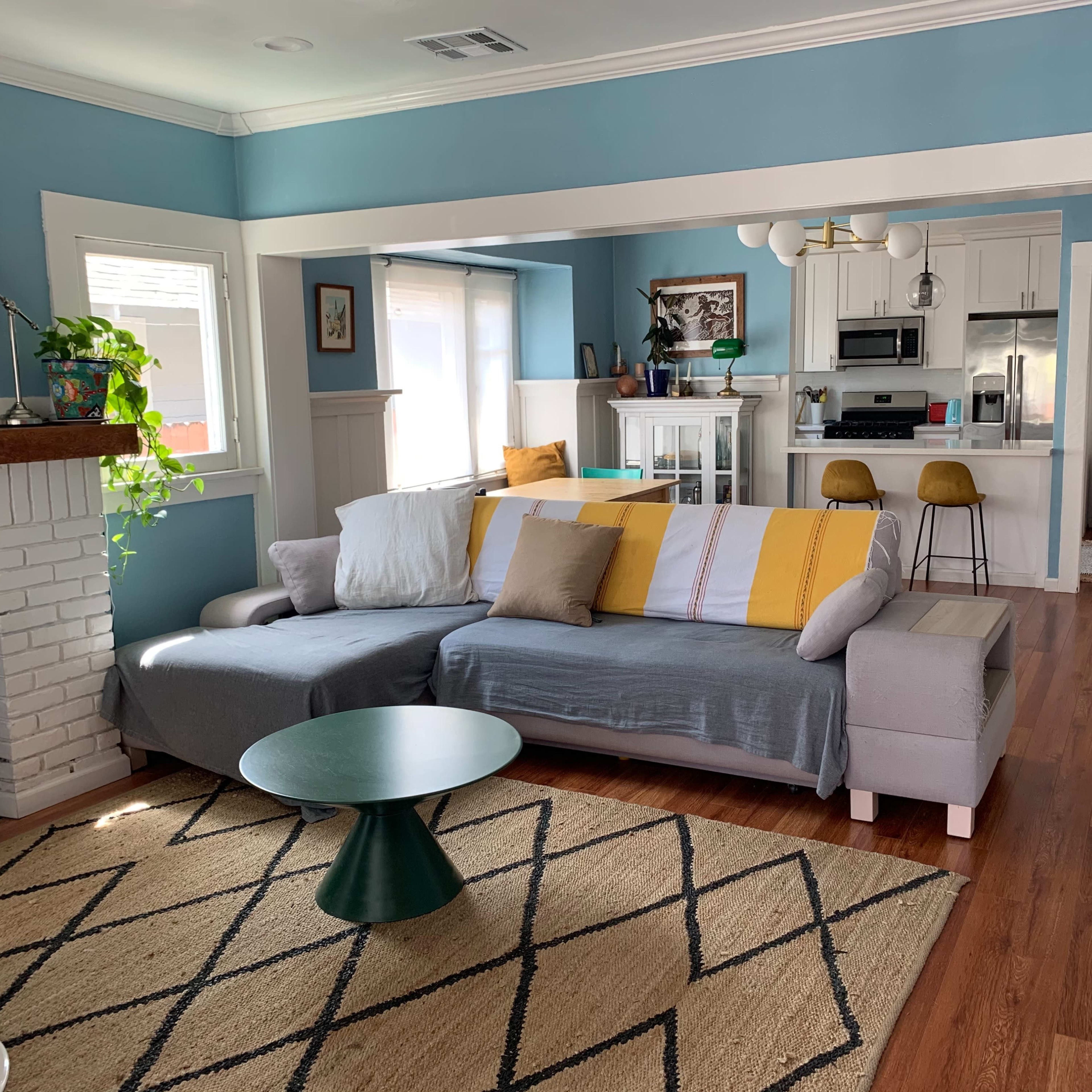 A living room with a sectional couch, a coffee table, and a view of an adjacent kitchen, featuring light wood flooring and pastel-colored walls.
