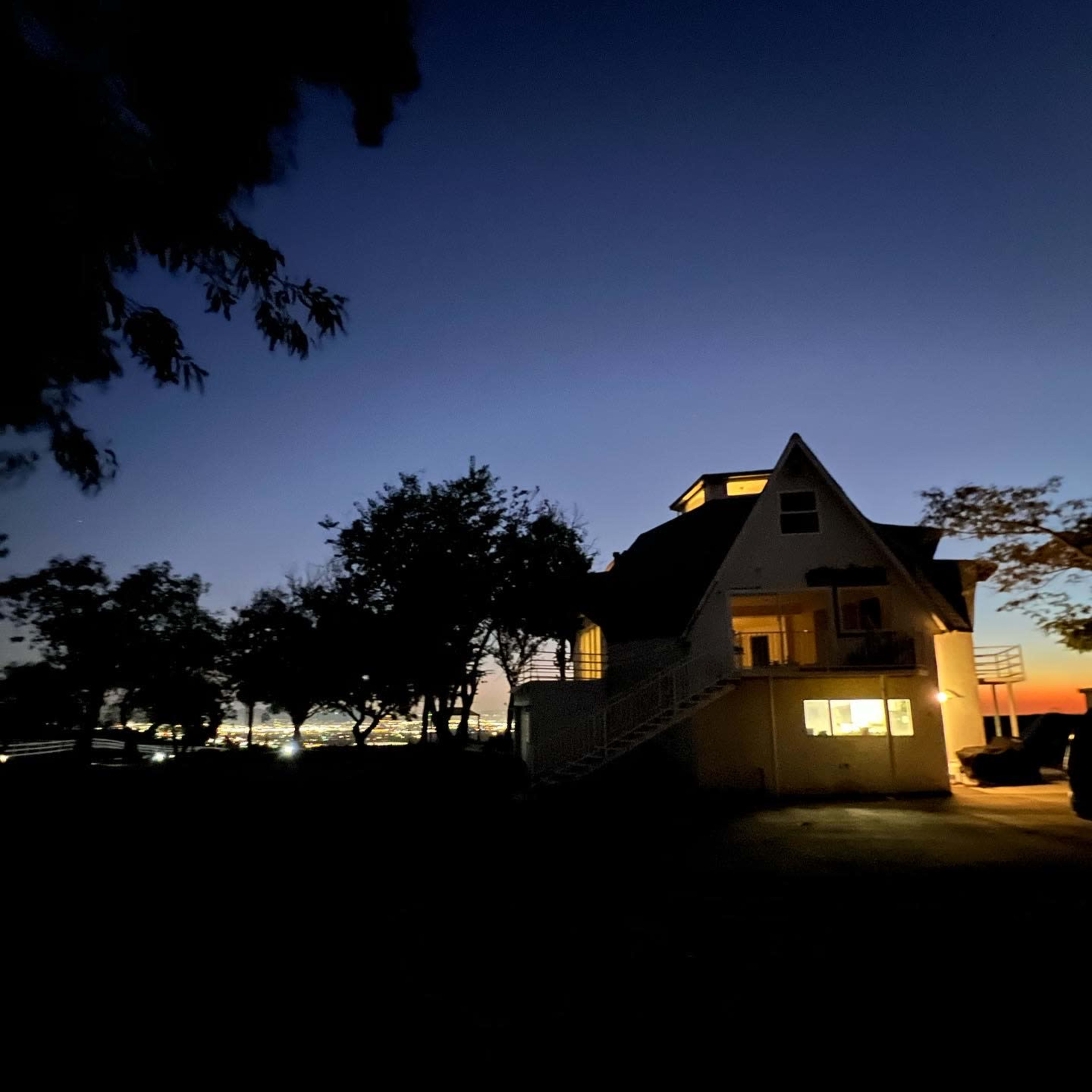A house with a triangular roof is illuminated at dusk, surrounded by trees against a darkening sky.