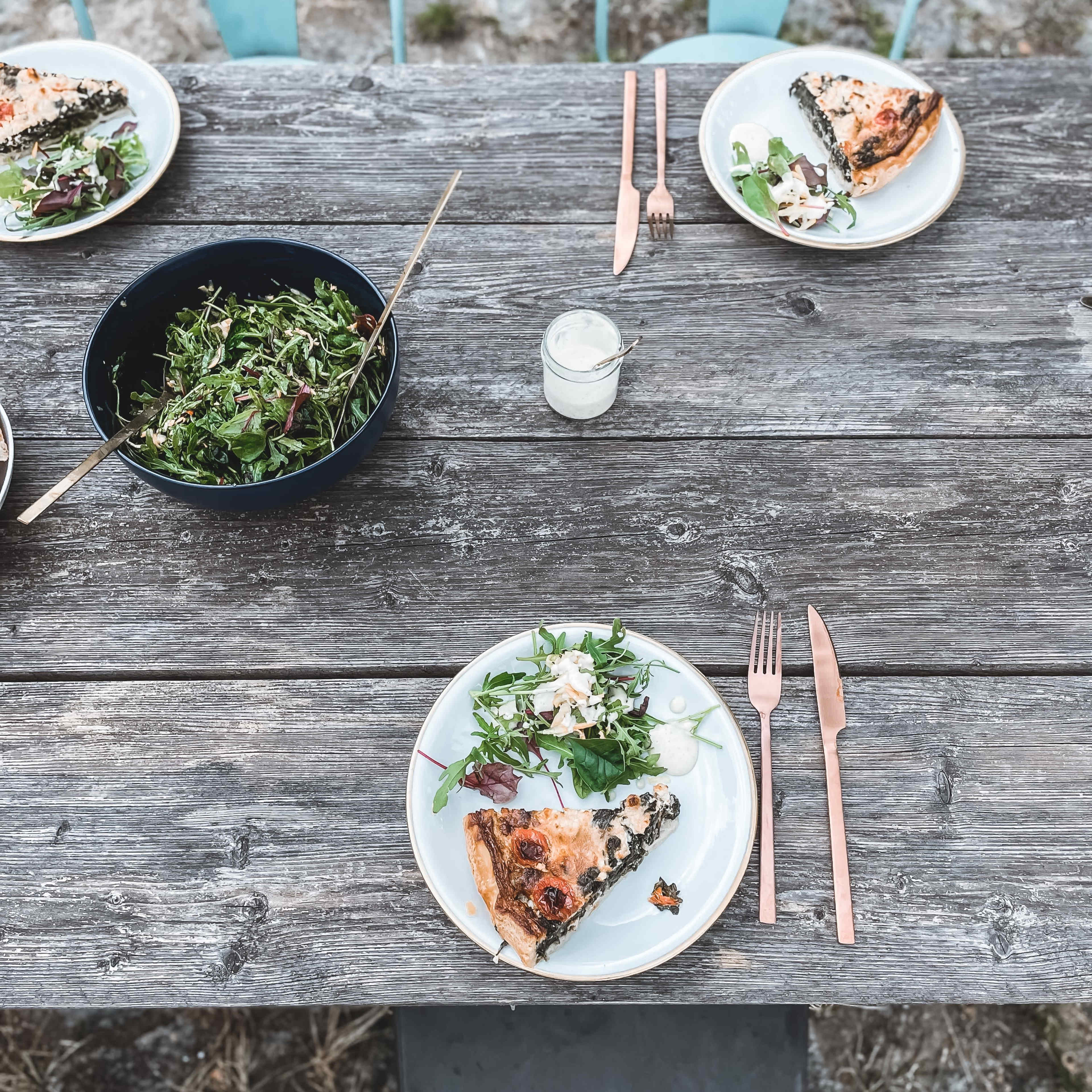 A rustic wooden table is set with plates of pizza and salads, accompanied by a bowl of greens and a small glass of dressing.