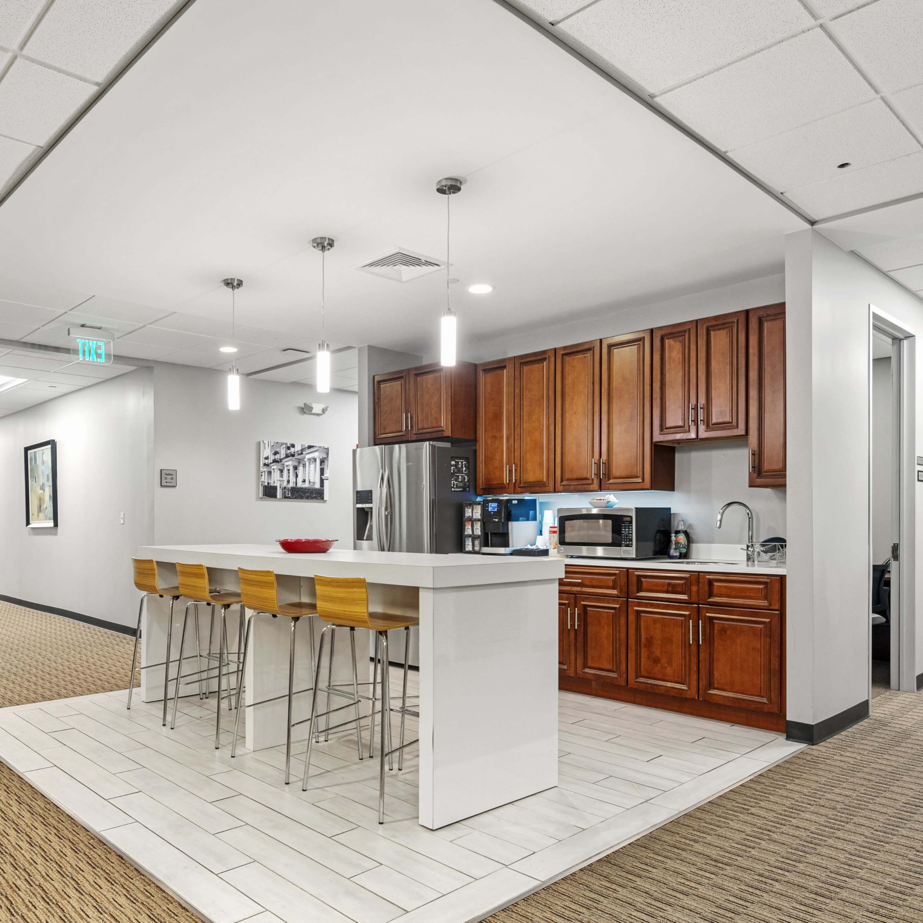 A modern kitchen area with a bar countertop and high stools, featuring wooden cabinets, a refrigerator, and a microwave.