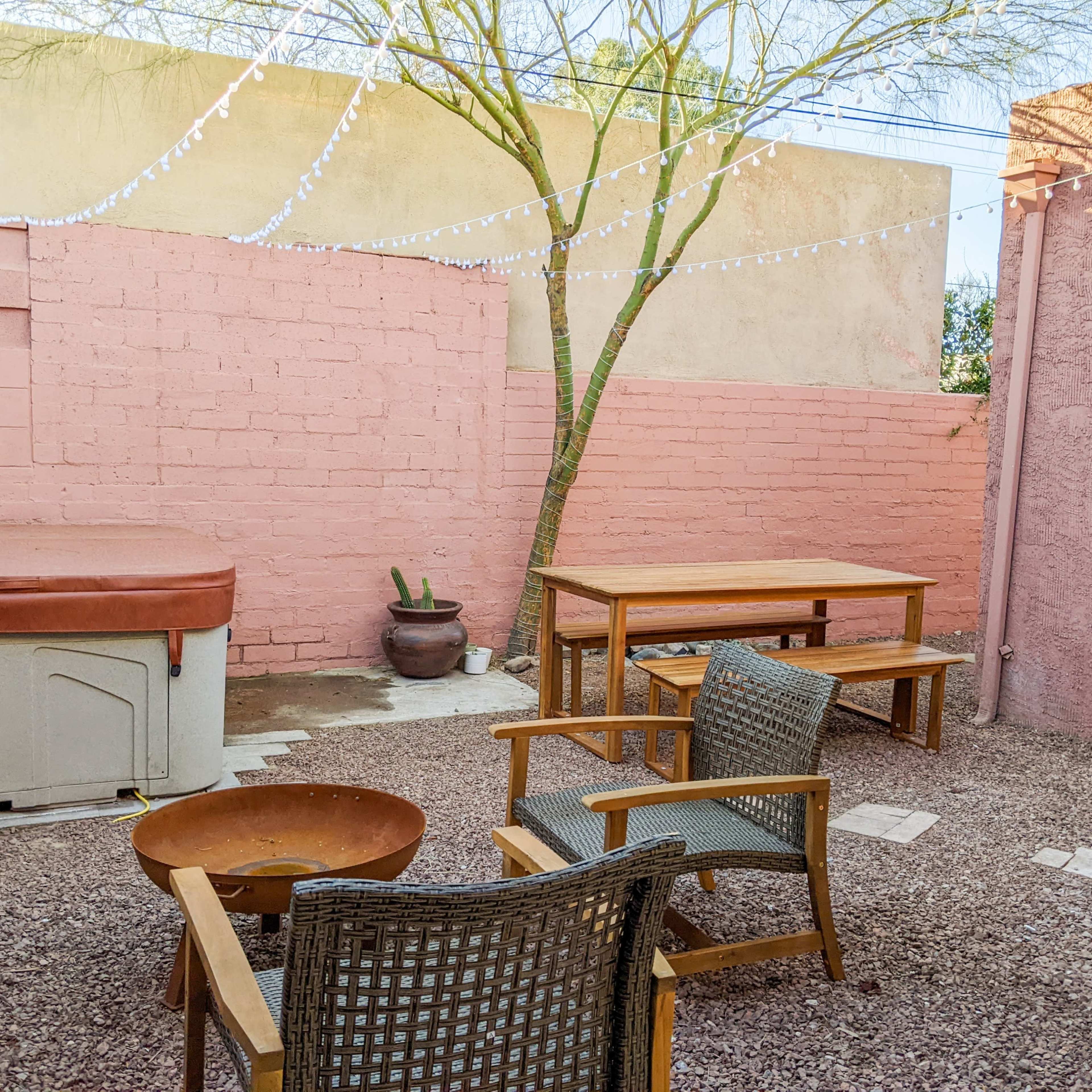 A small outdoor patio area featuring a hot tub, a wooden dining table, four chairs, and a fire pit, surrounded by pink walls and a tree.