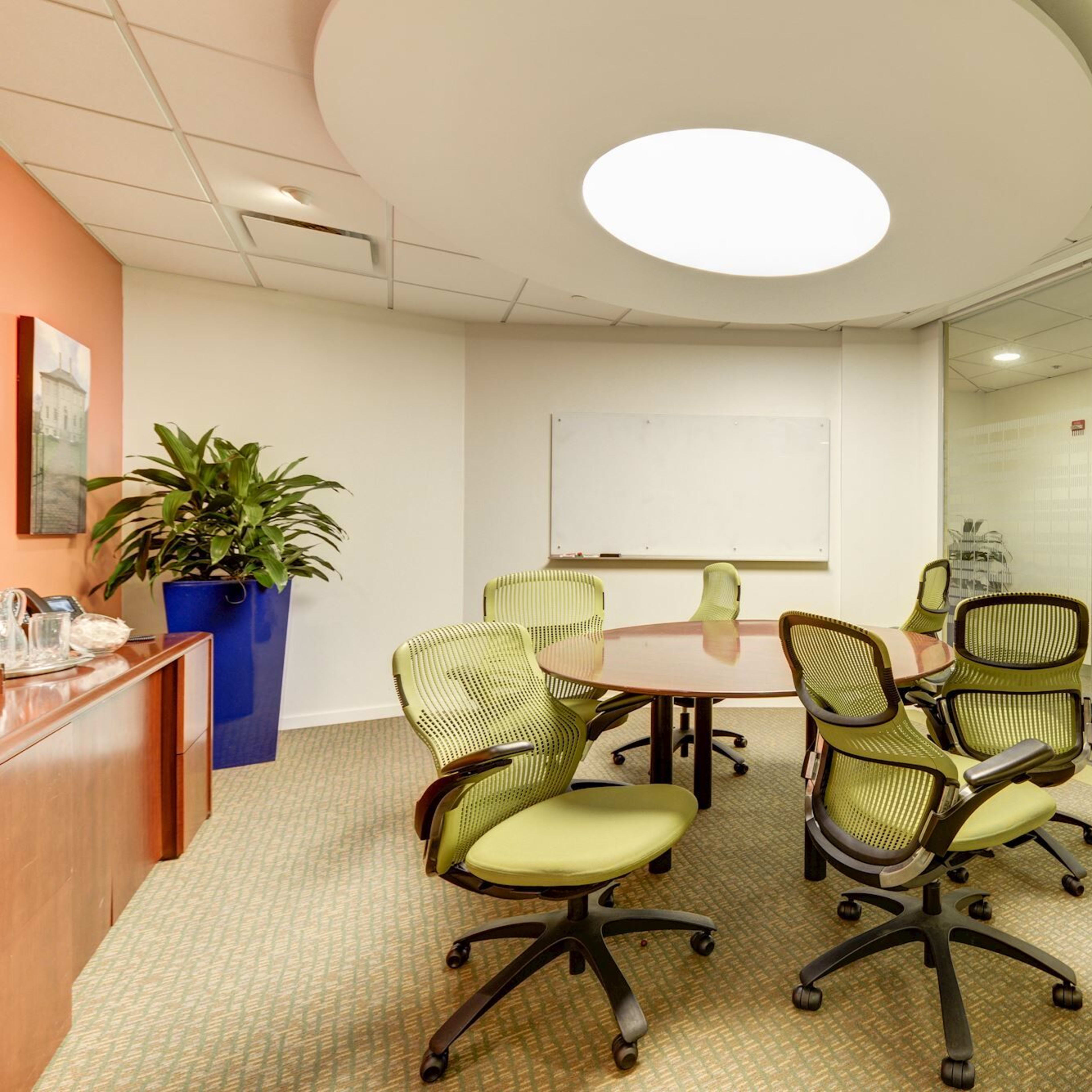 A conference room features a round table surrounded by green chairs, with a whiteboard and glass walls.