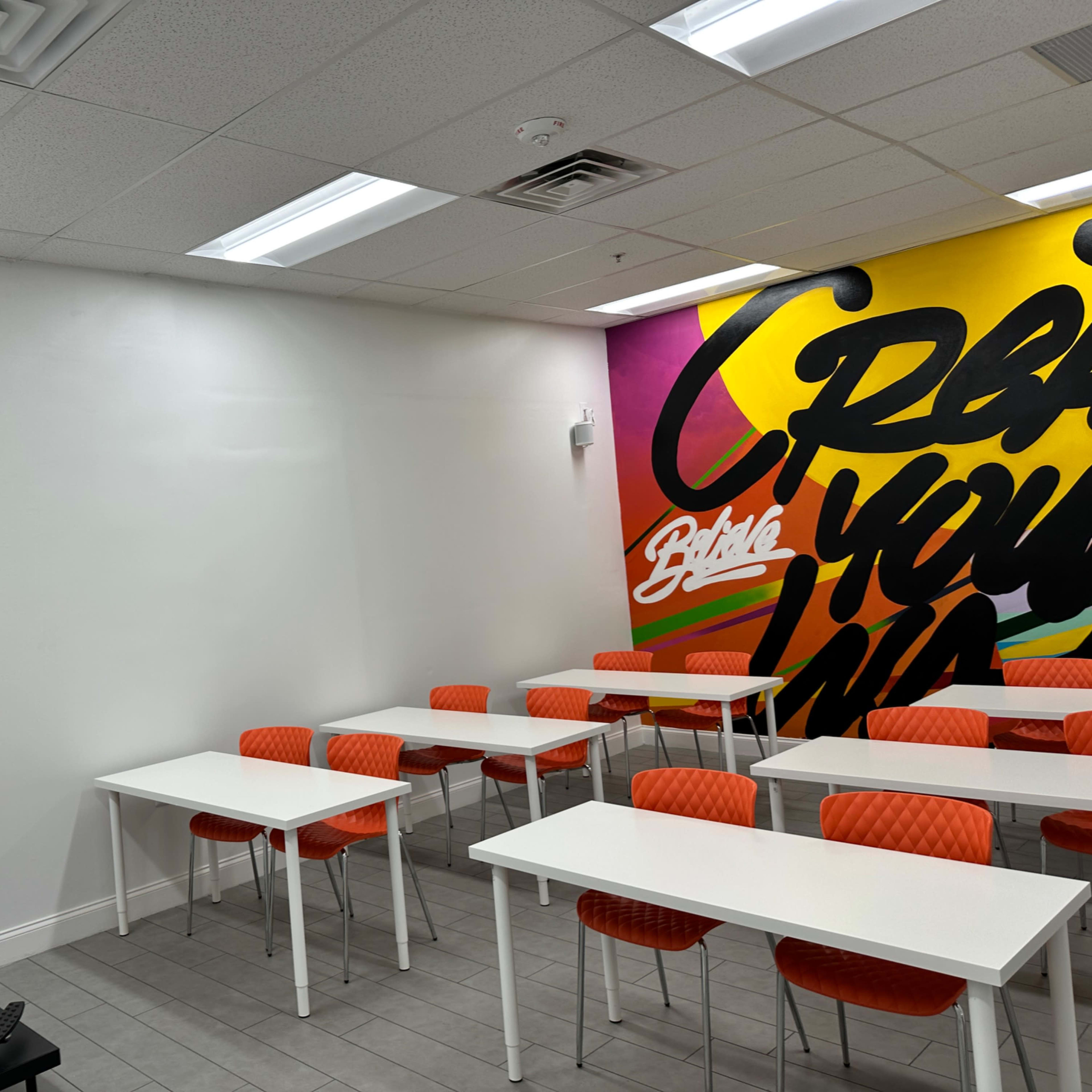 A brightly colored classroom with a white styling chair on one side and rows of white tables with orange chairs.