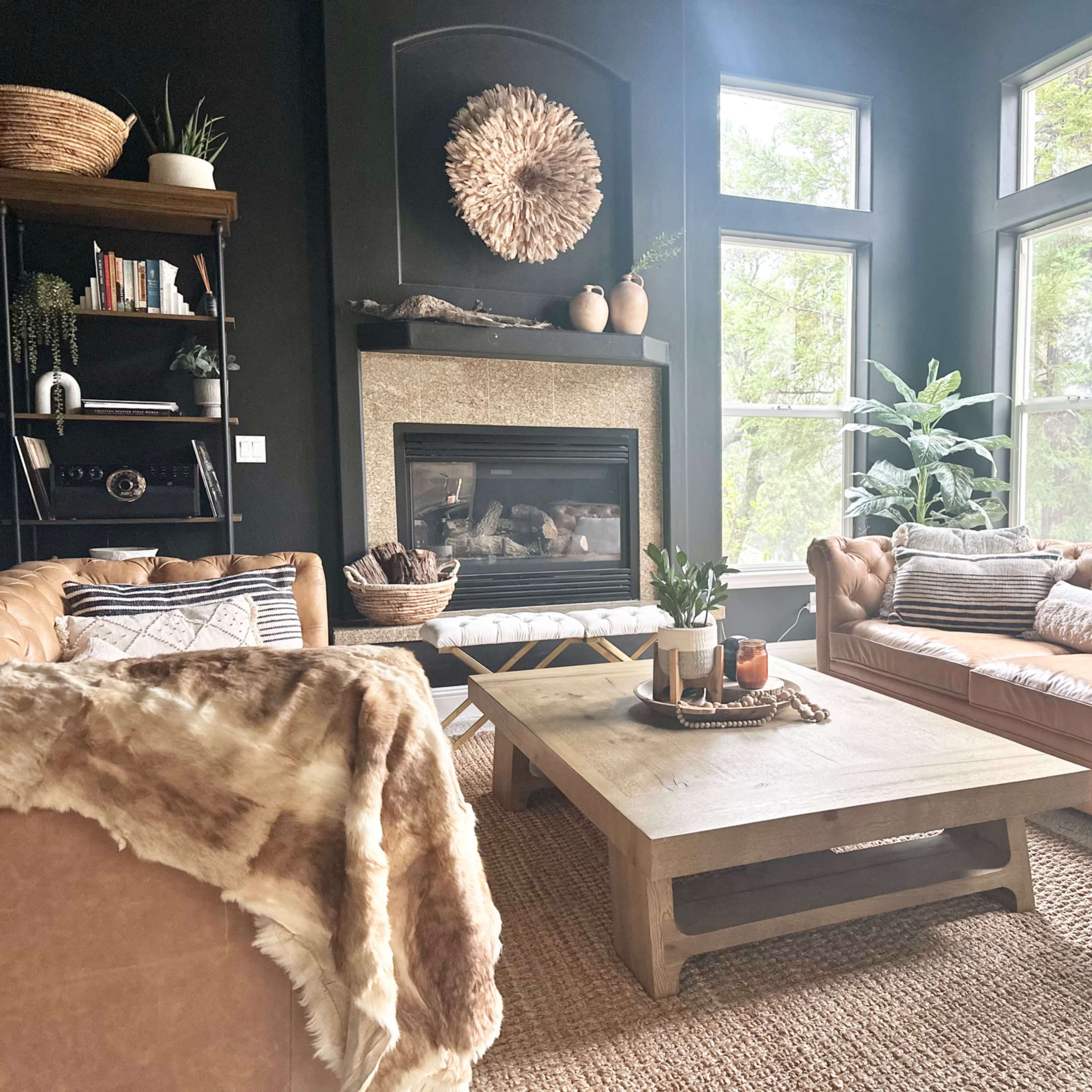 A cozy living room features a stone fireplace surrounded by large windows, two leather sofas, a wooden coffee table, and decorative plants.