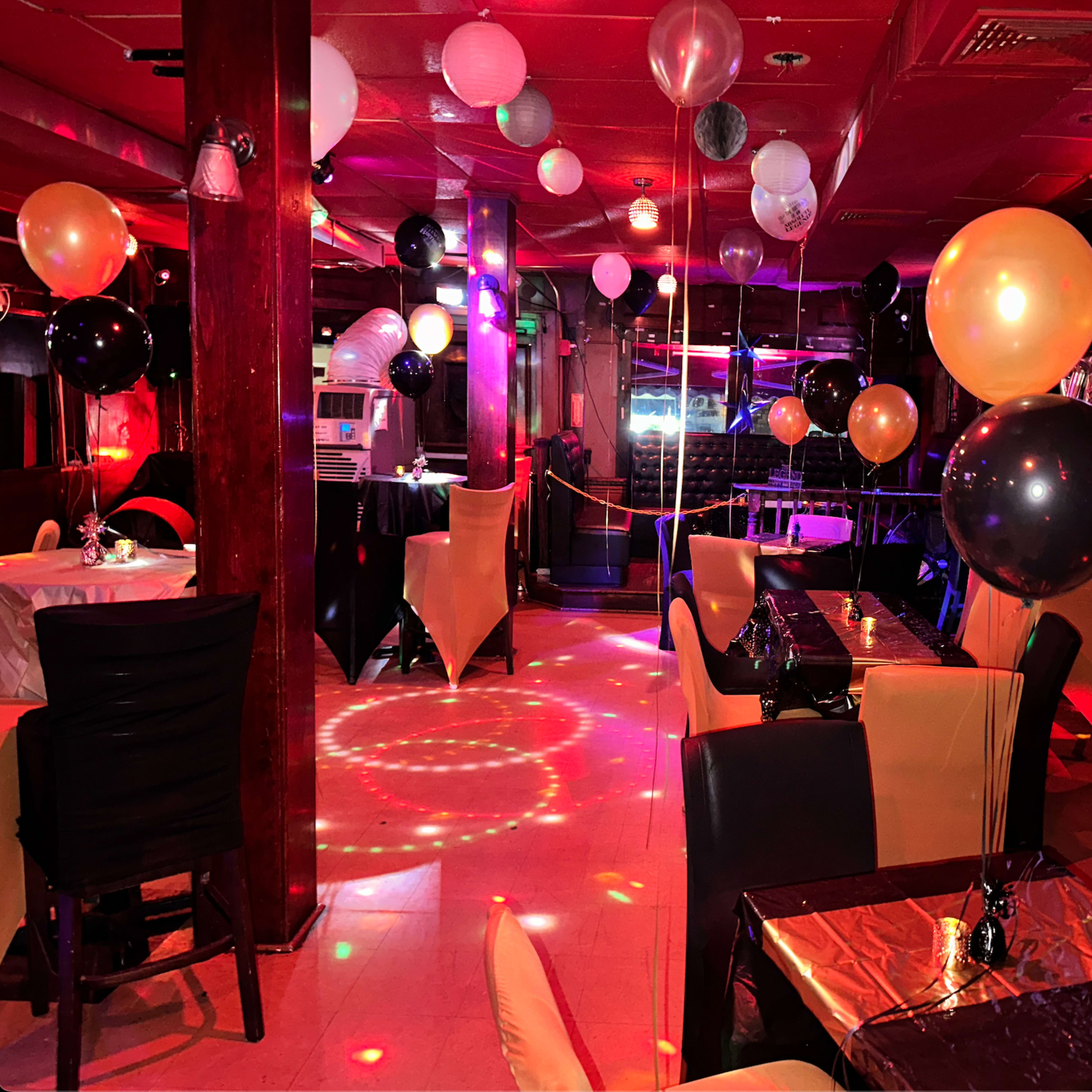 The interior of a party venue is decorated with black, gold, and white balloons, featuring tables set for guests under colorful lights.