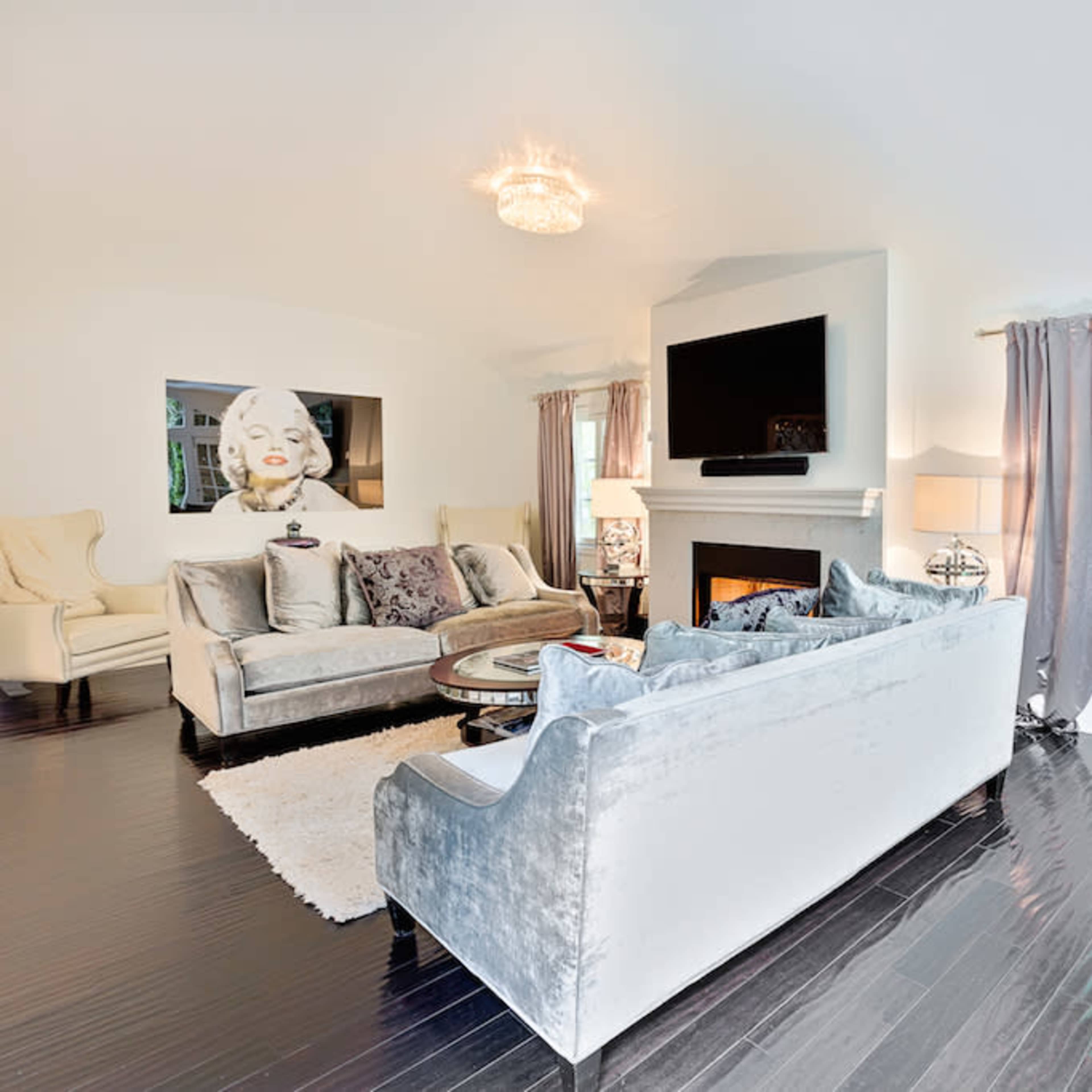 The living room features a sleek sectional sofa, a circular coffee table, and a large flat-screen TV mounted above a fireplace, complemented by elegant curtains and decor.