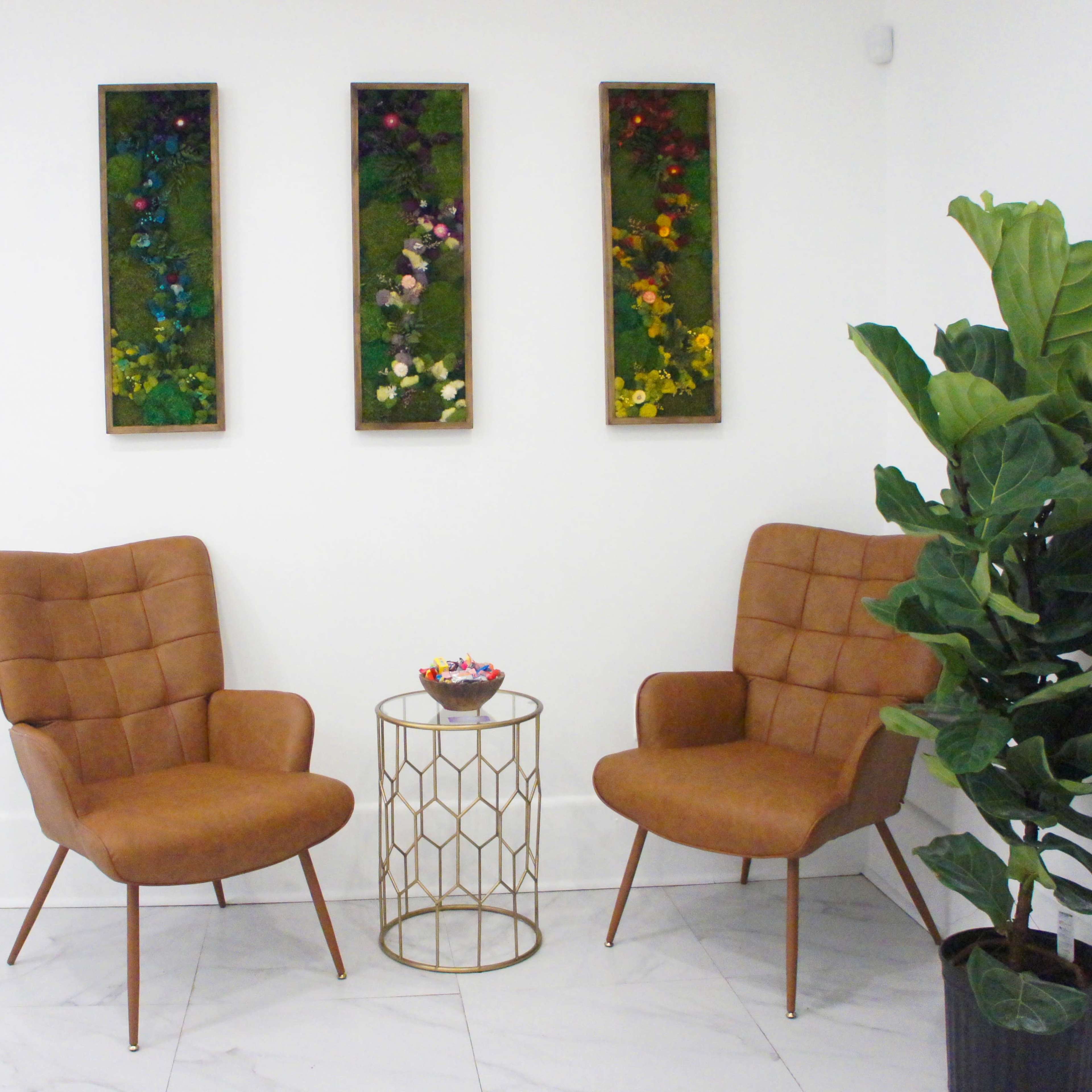 Two brown upholstered chairs sit opposite each other with a gold geometric side table between them, alongside a large potted plant and three framed pieces of floral wall art.