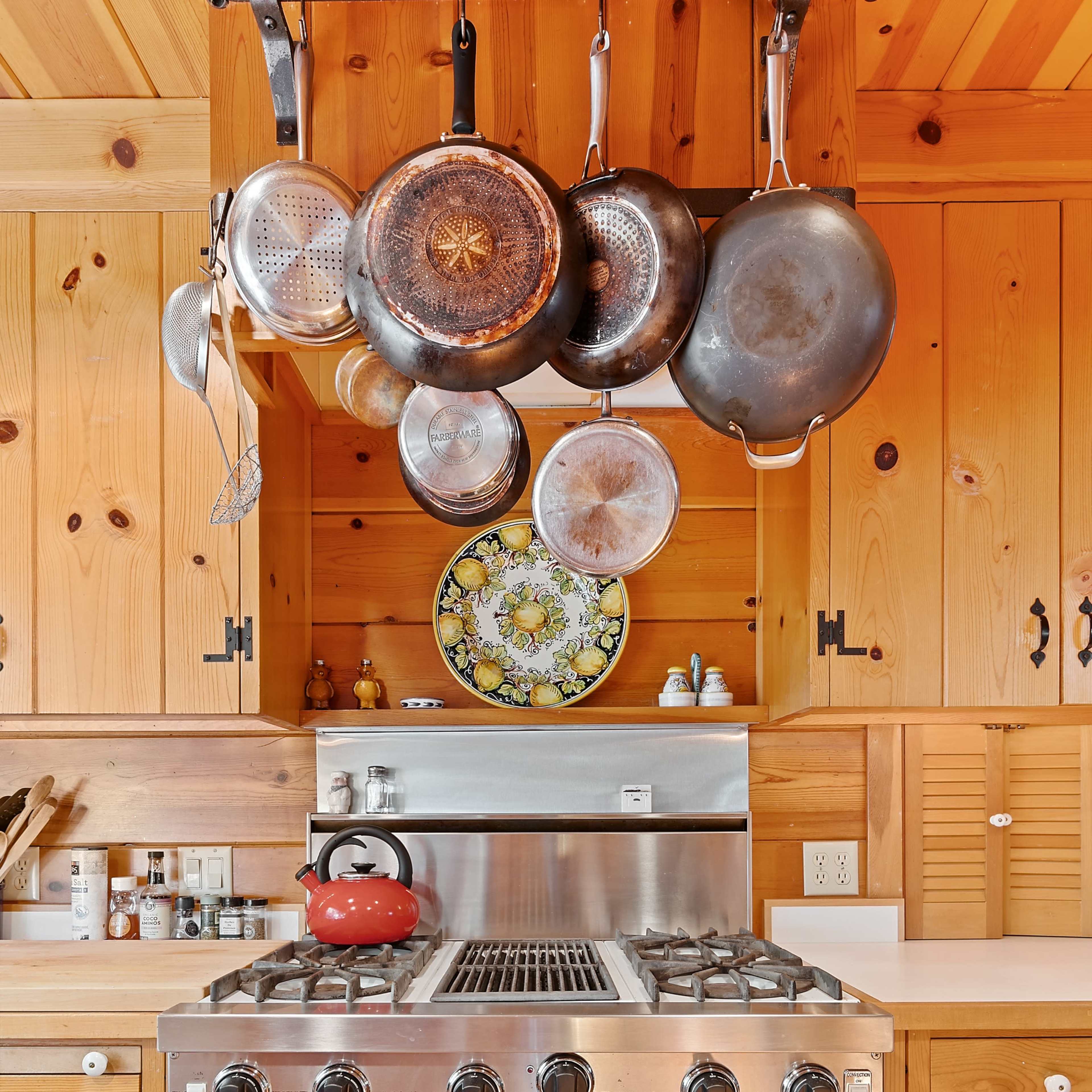 A stainless steel Viking gas range is centered in a wooden kitchen with hanging cookware above it and a red kettle.