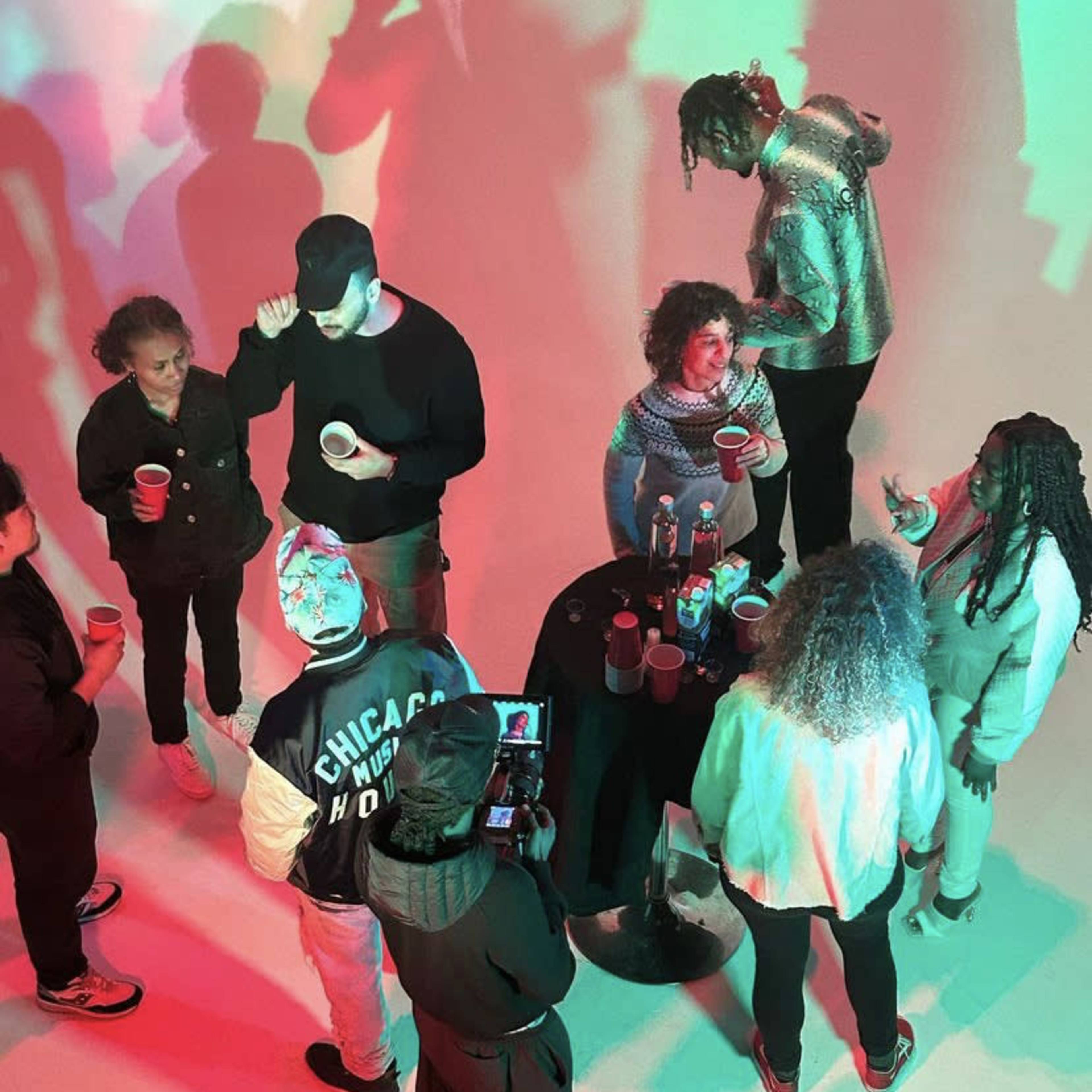 A group of people gathers around a table with red cups and drinks in a brightly lit, multi-colored setting.