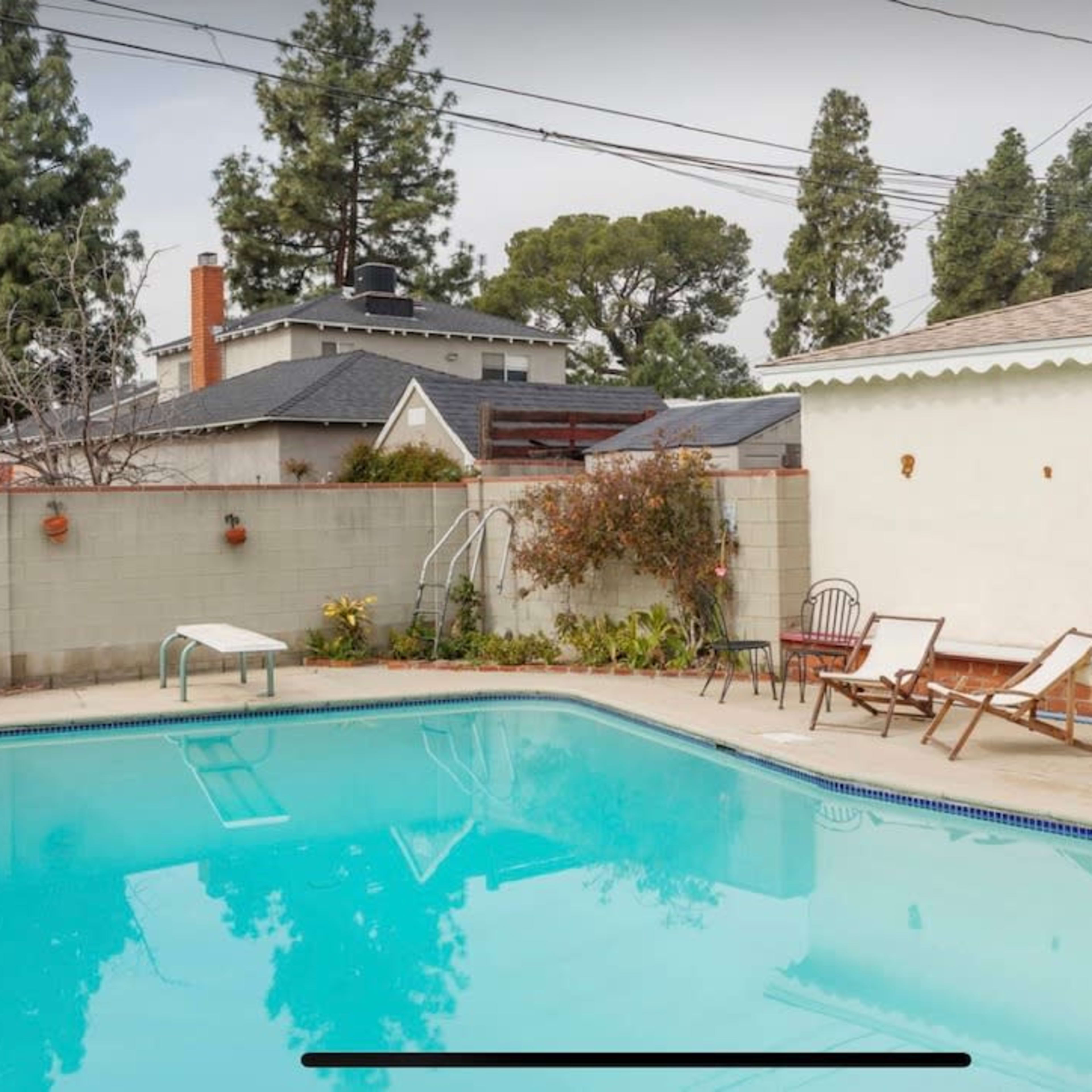 A swimming pool surrounded by concrete and greenery, with two lounge chairs positioned nearby.