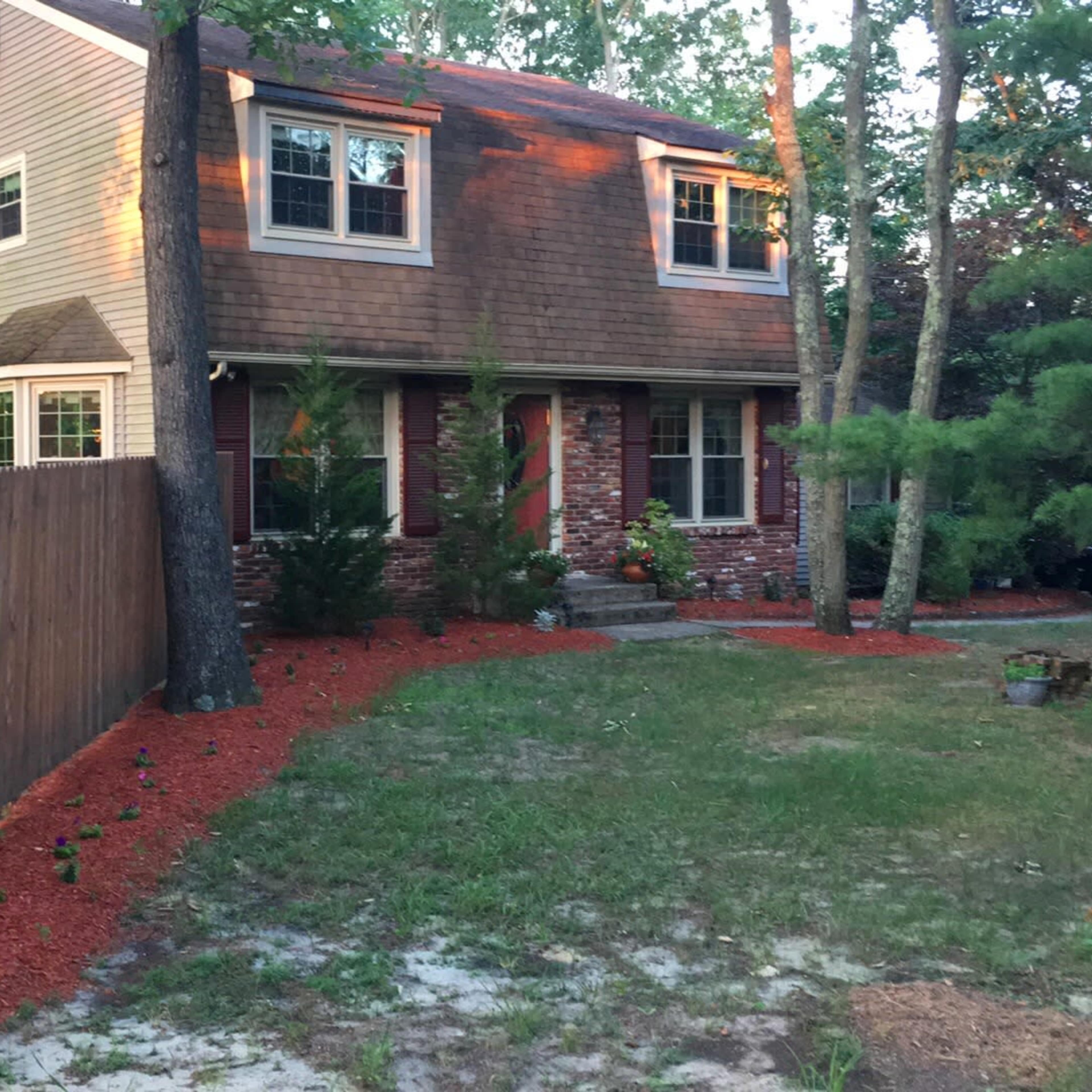 A two-story brick house surrounded by trees, with red mulch covering the landscaping and a wooden fence on one side.