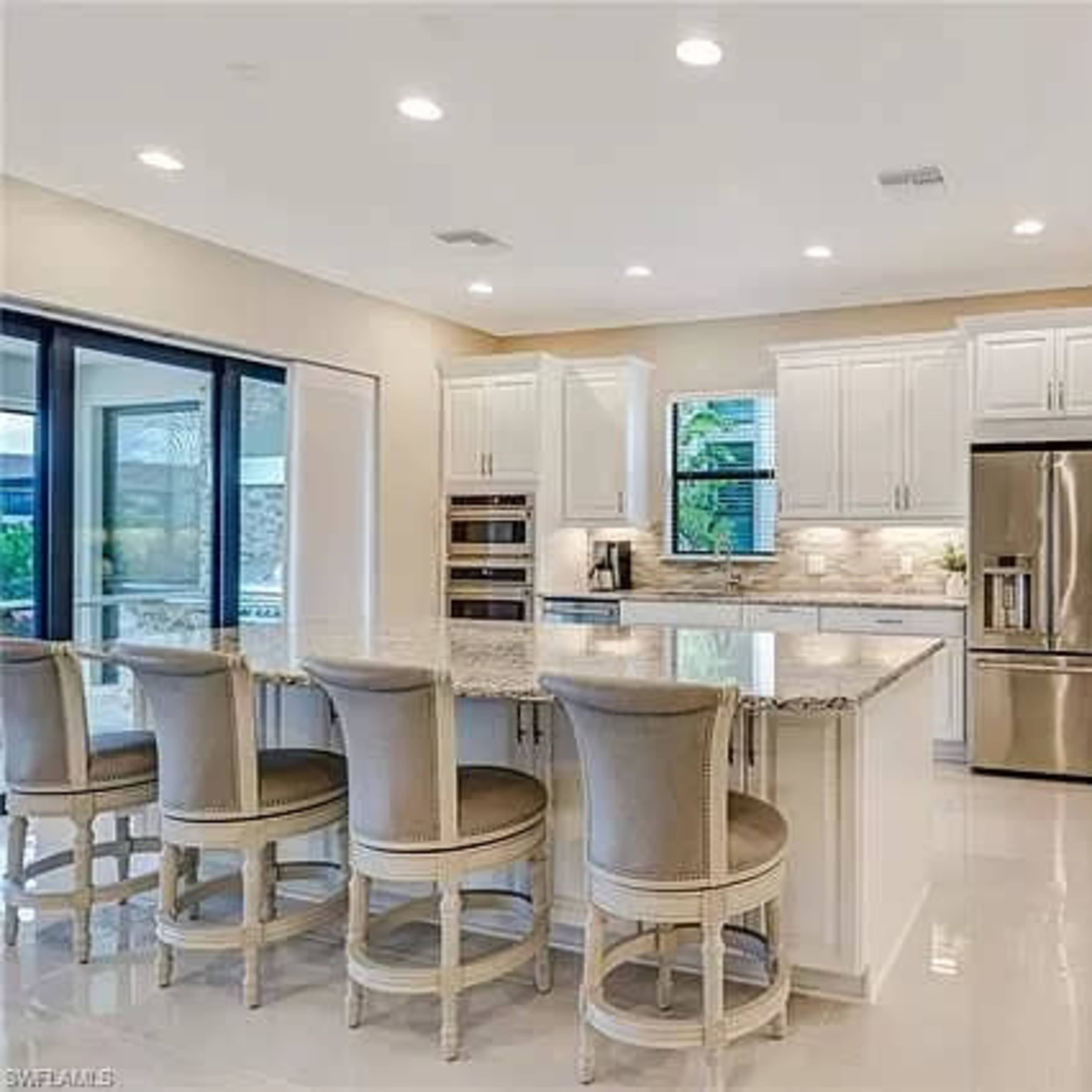 A modern kitchen features white cabinetry, a large island with bar seating, stainless steel appliances, and natural light from large windows.
