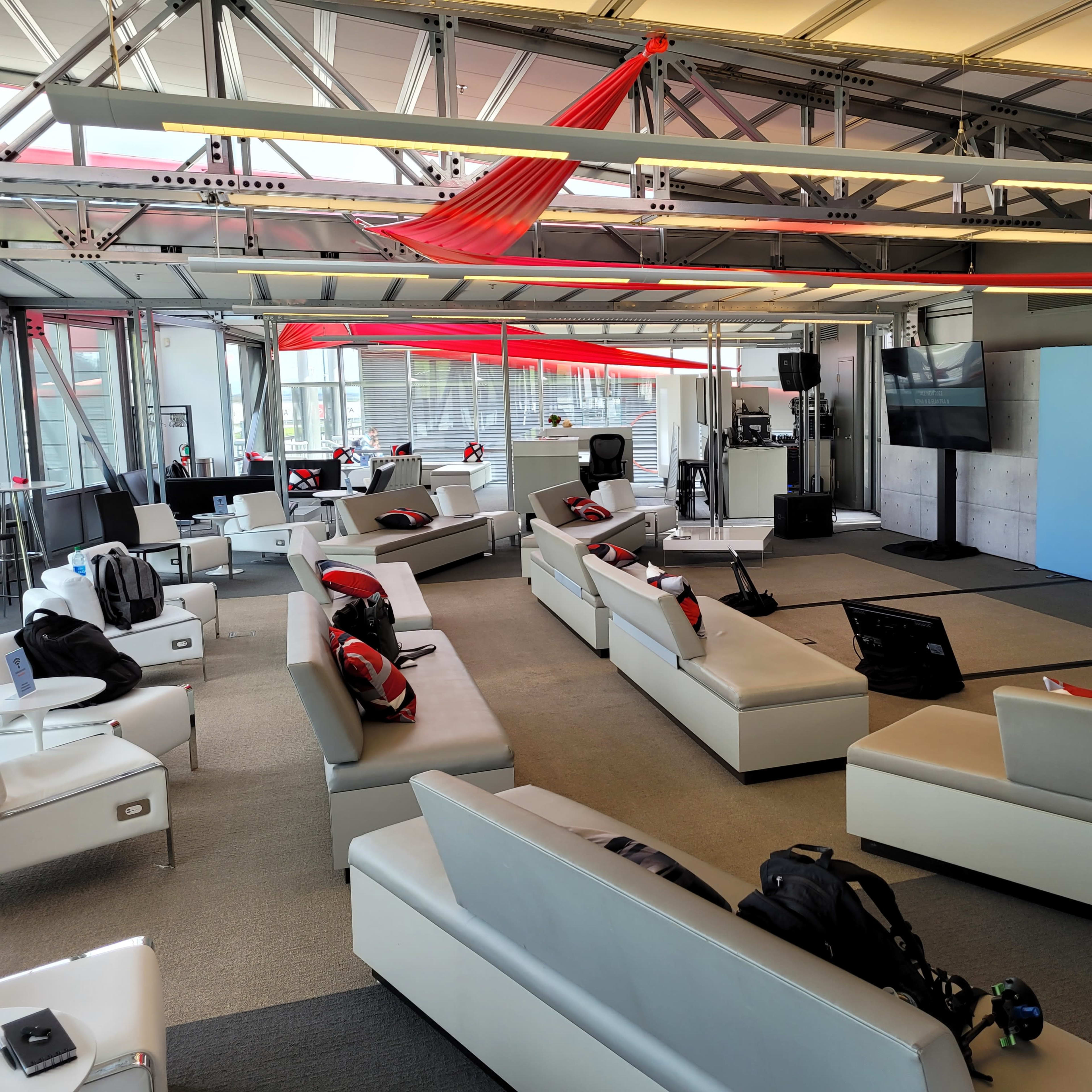 A modern, open lounge area furnished with white sofas, equipped with red and black accent pillows, and featuring a blue backdrop display with a logo.